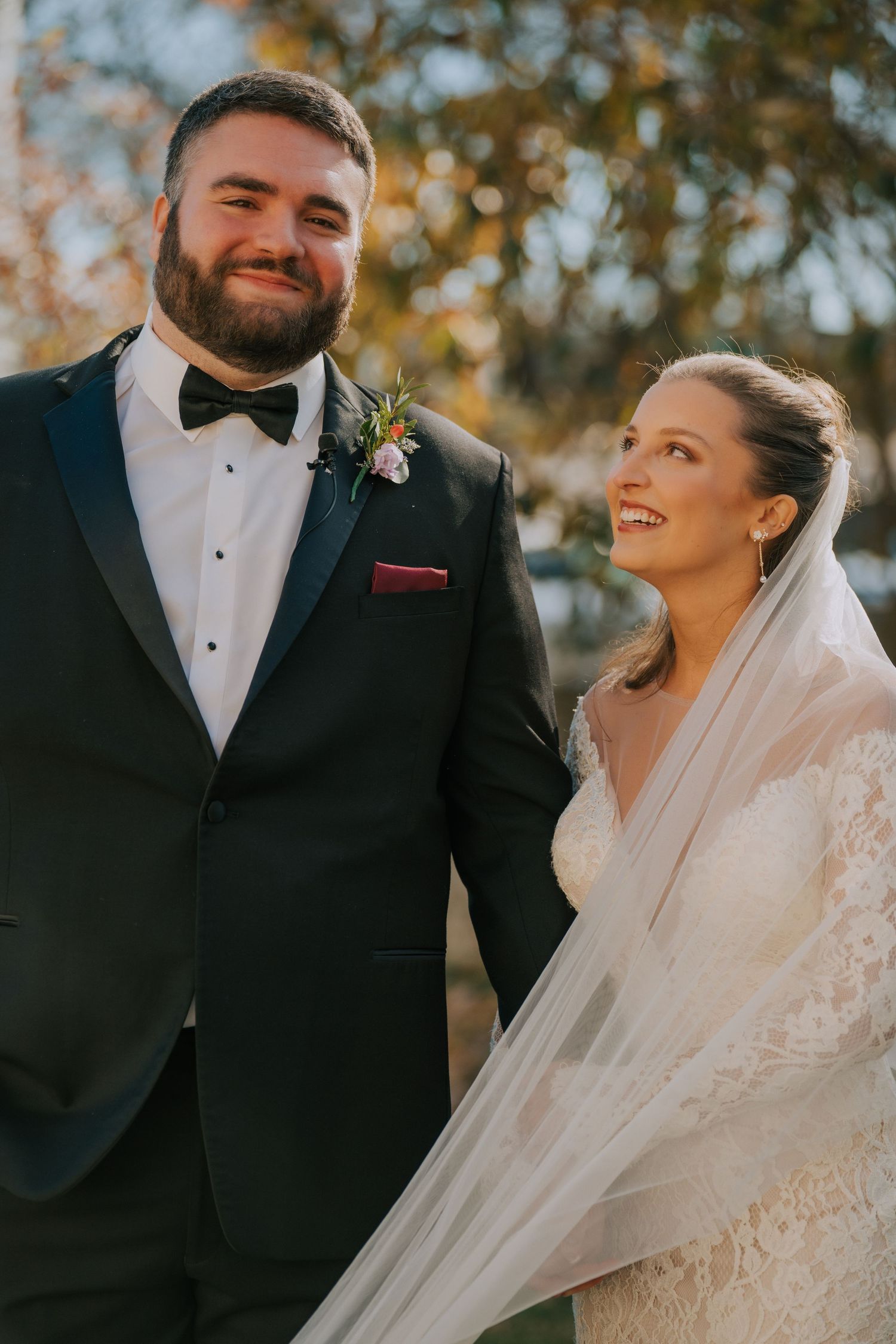 A joyful wedding couple shares a candid moment outdoors during golden hour, with autumn foliage in the background.