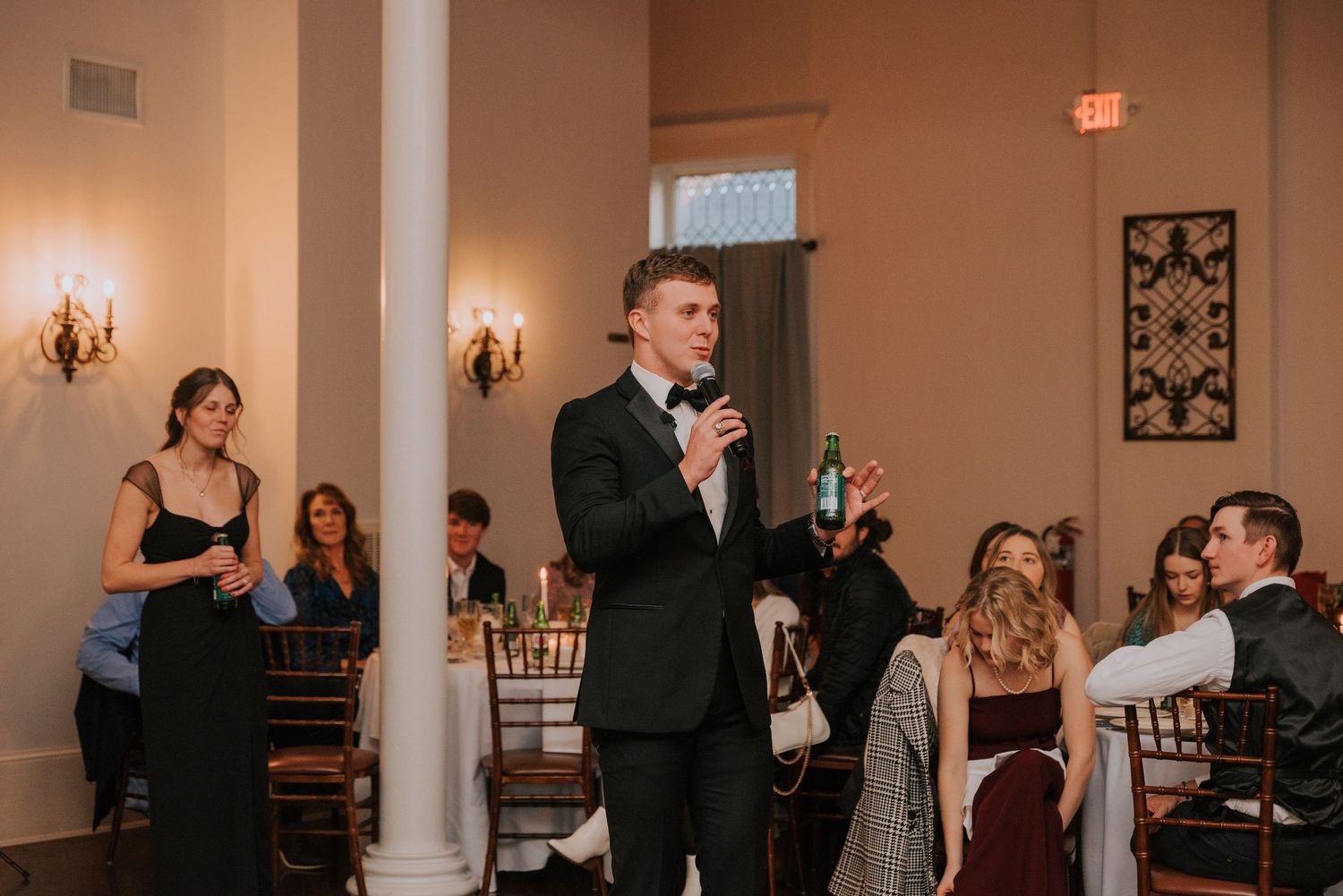 A person in a black tuxedo gives a speech at an elegant indoor wedding reception with warm lighting.
