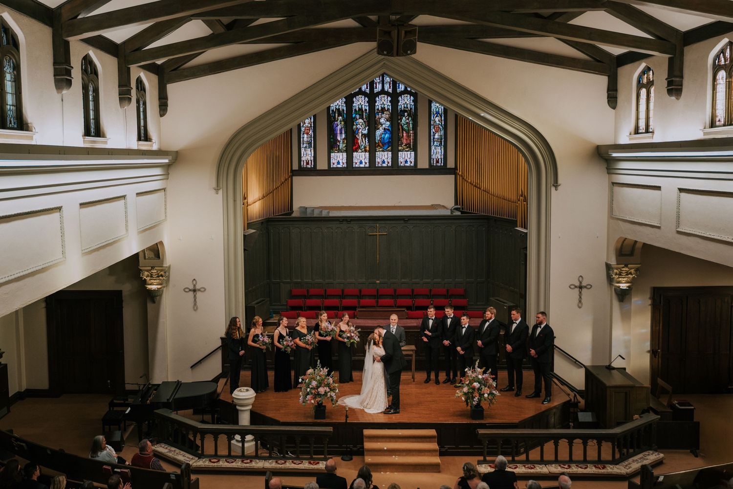 A choir performs on stage in a traditional church sanctuary with stained glass windows and wooden beams overhead.