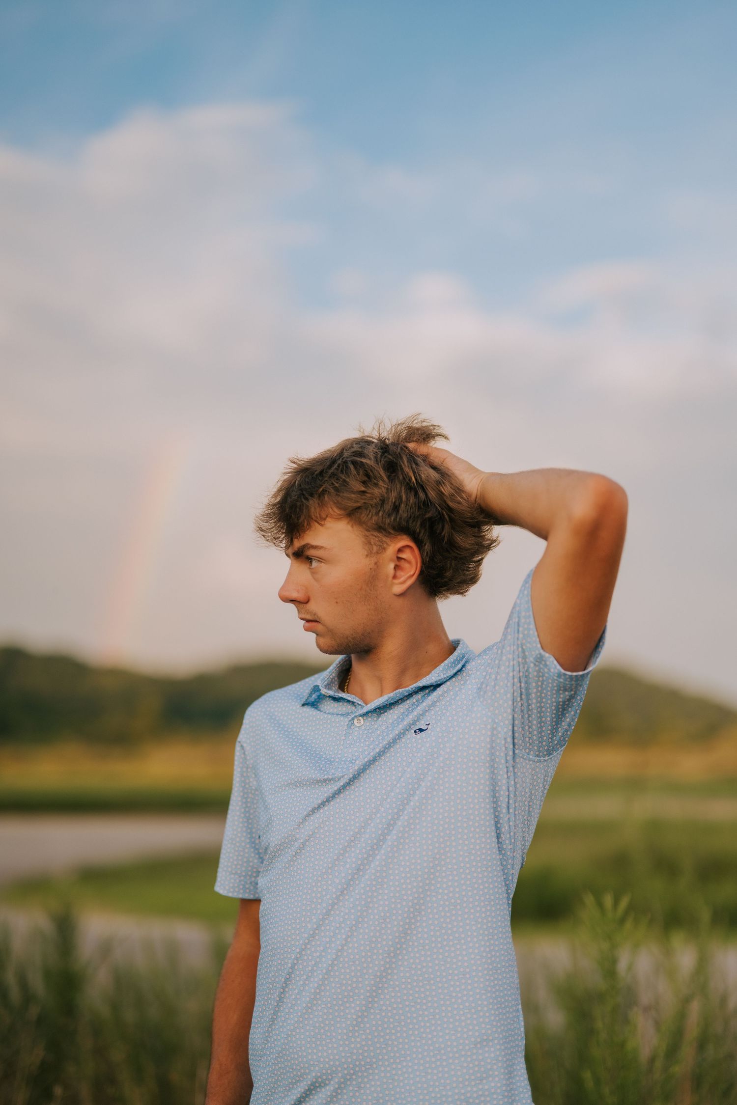 Person in light blue polo shirt poses against scenic rural landscape at sunset.