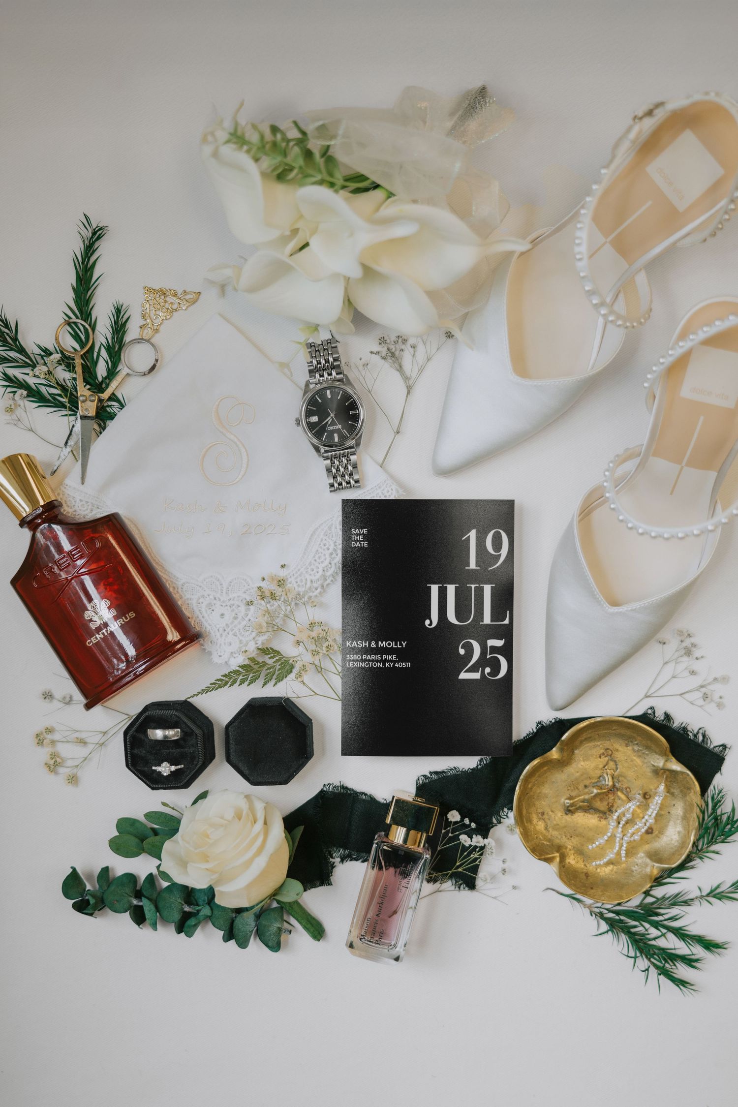 Wedding details flatlay with white shoes, perfume bottles, and greenery on light background.