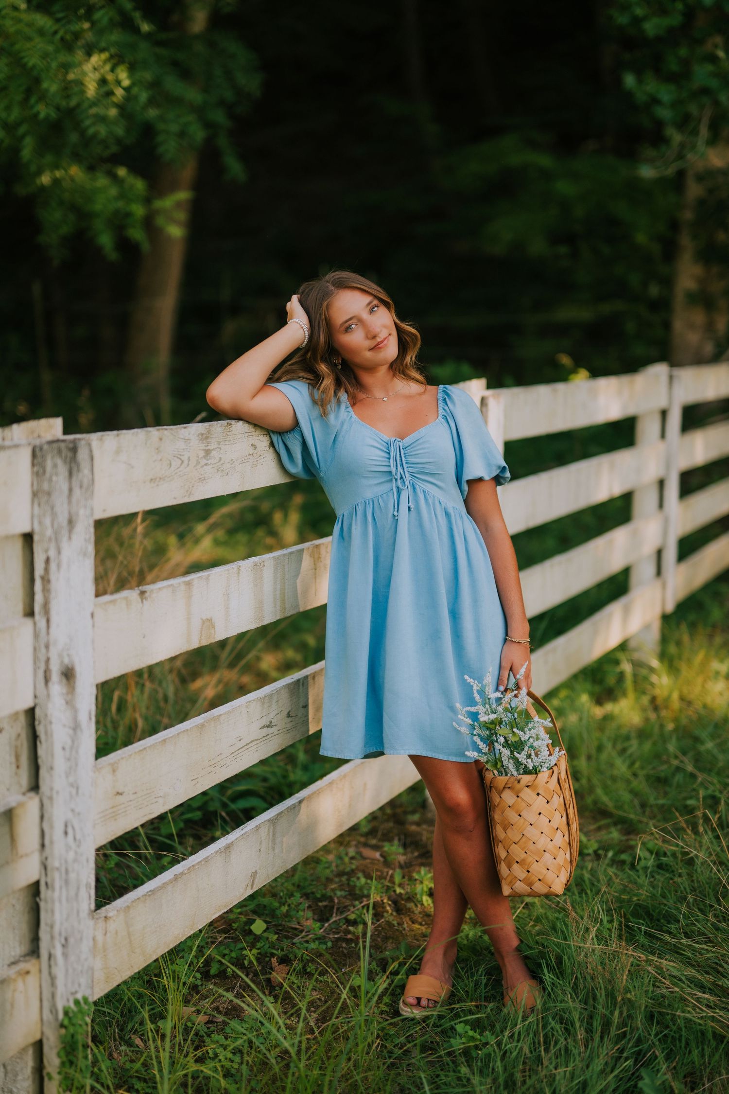 Person in blue dress poses playfully along white fence in late afternoon sunlight.