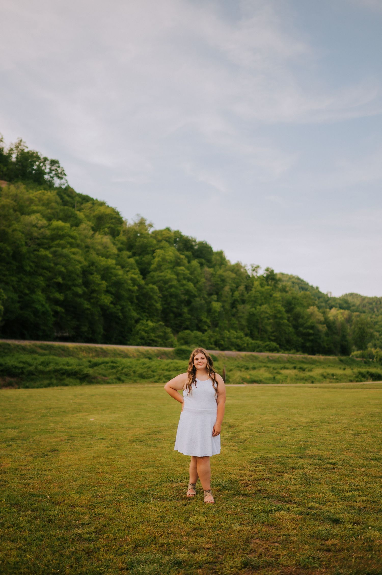 A person in white stands in an open green field with mountains in the background.