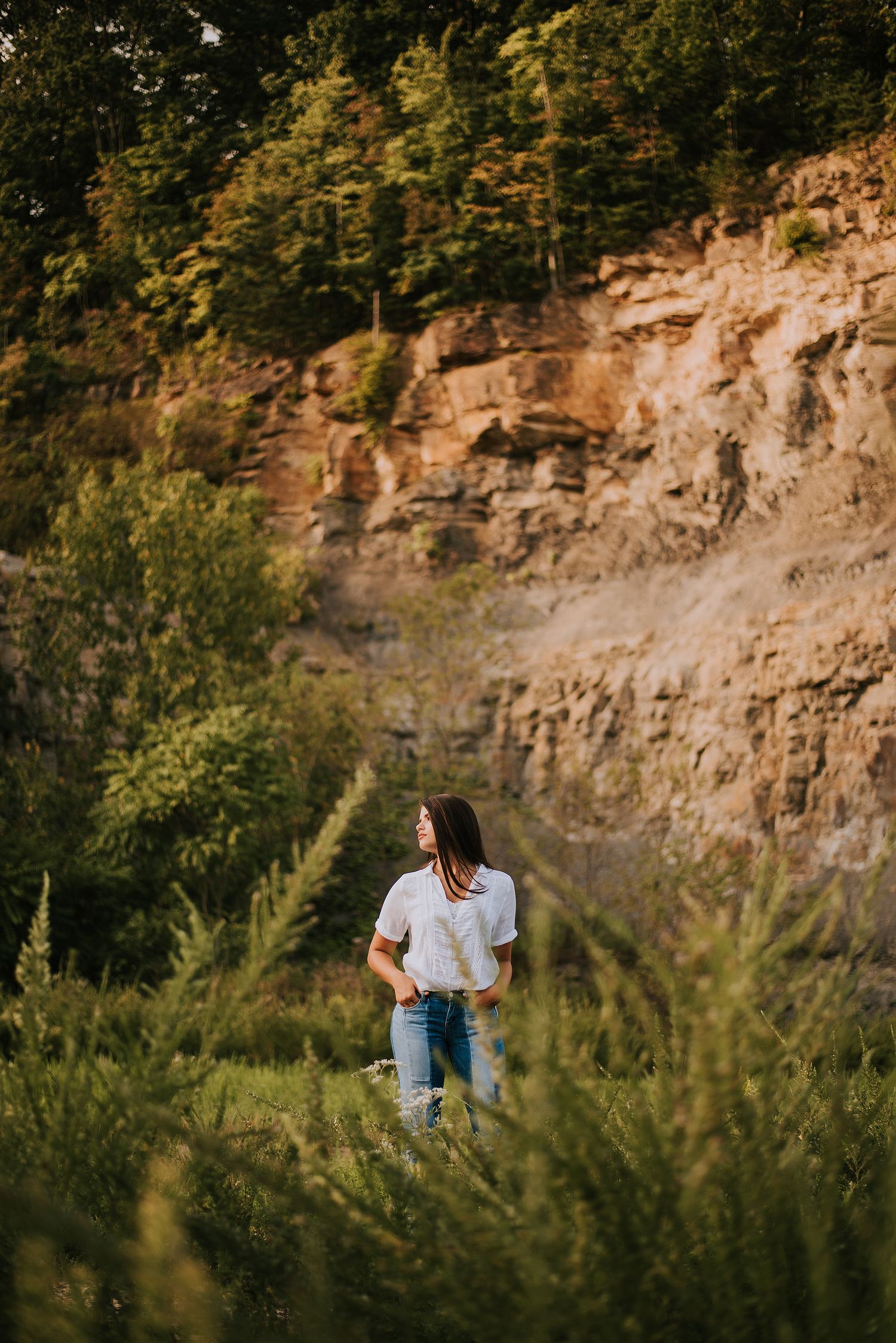 A person in casual attire stands near a rocky cliff face surrounded by lush greenery.