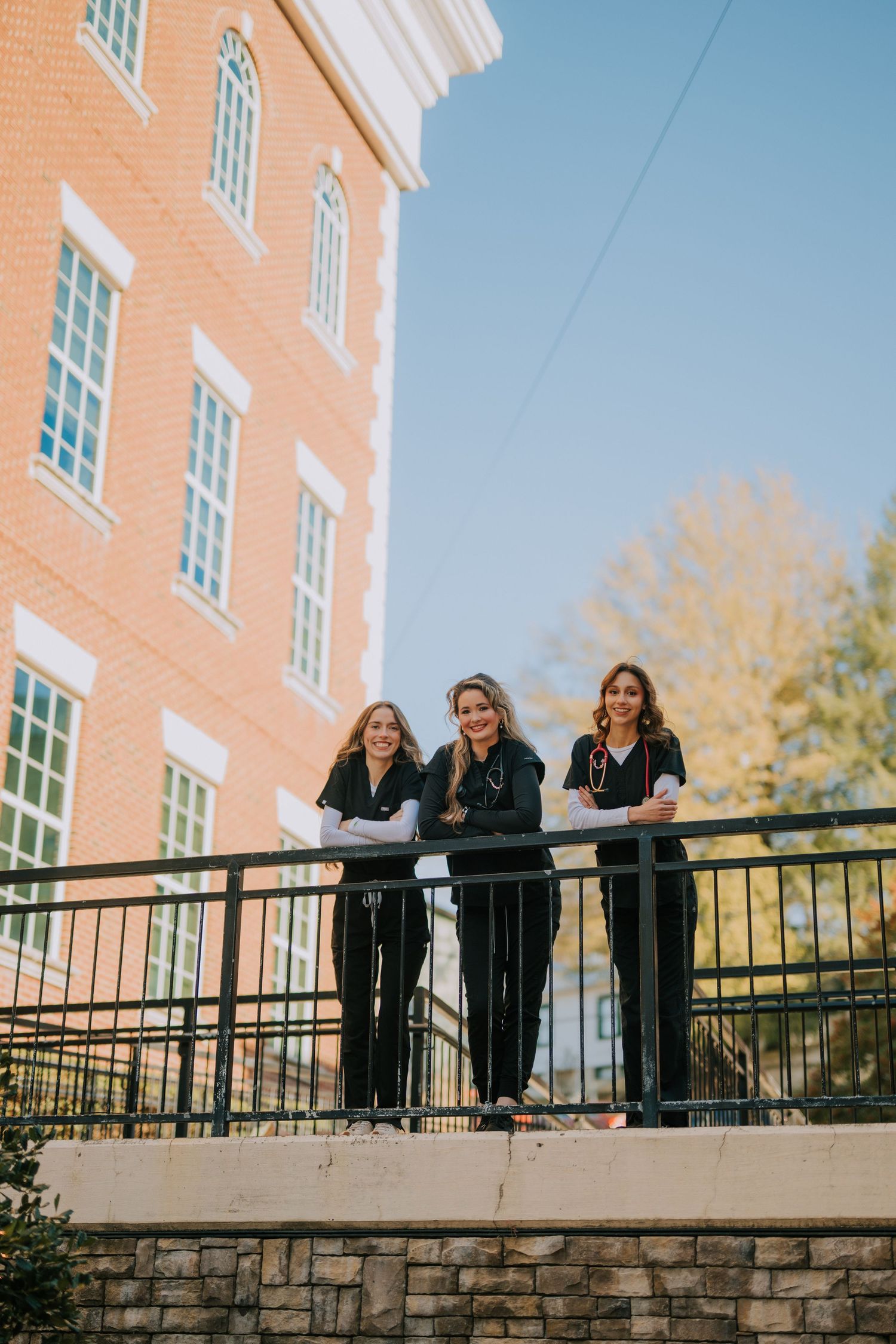 People in black attire pose along a stone wall railing of a brick building.