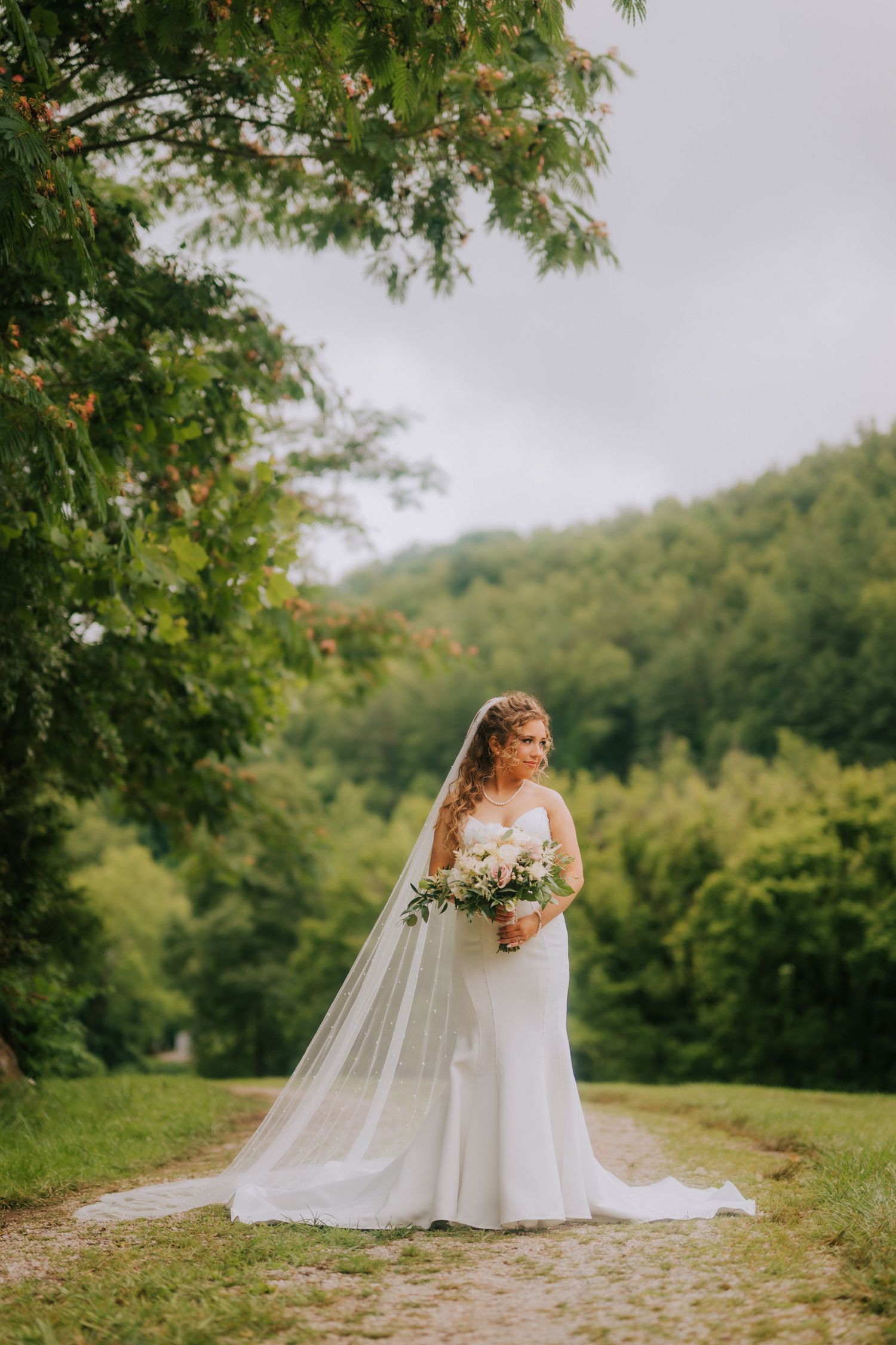 Bride in white gown and veil holds bouquet on garden path surrounded by lush green trees.