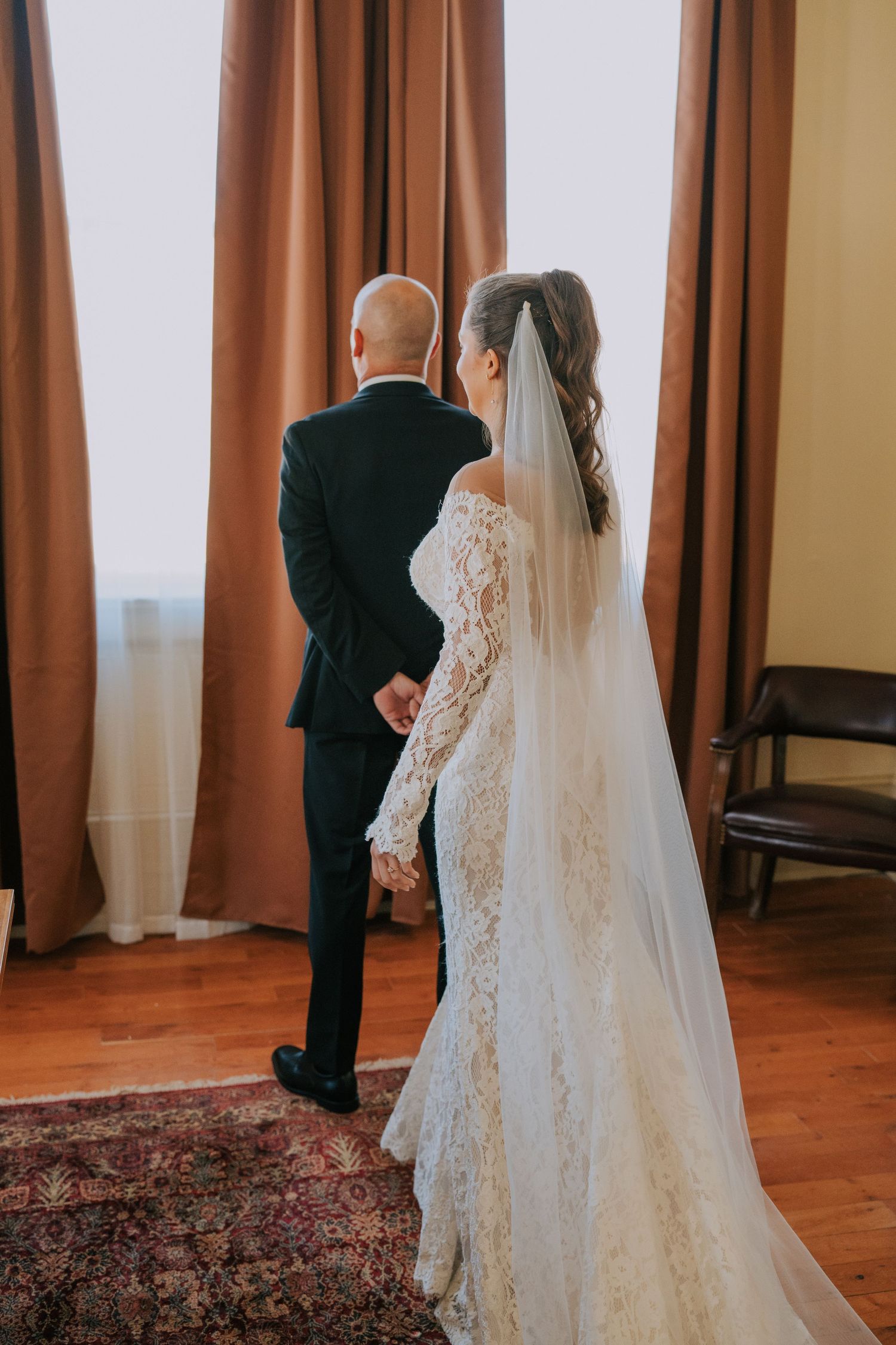 A bride in a lace wedding gown with long veil stands behind a man in a black suit near brown curtains.