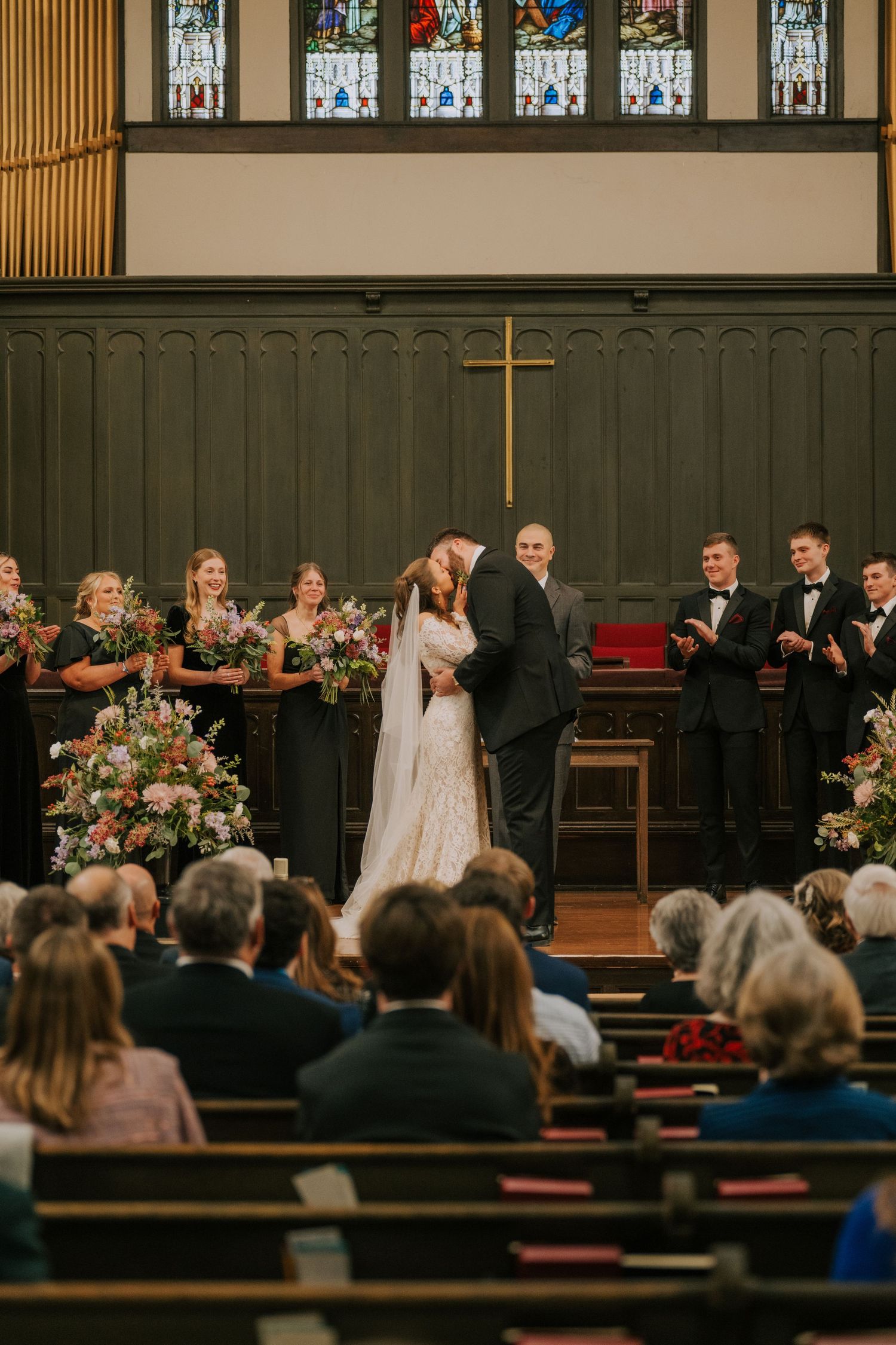 A traditional church wedding ceremony with stained glass windows, floral arrangements and gathered guests in wooden pews.