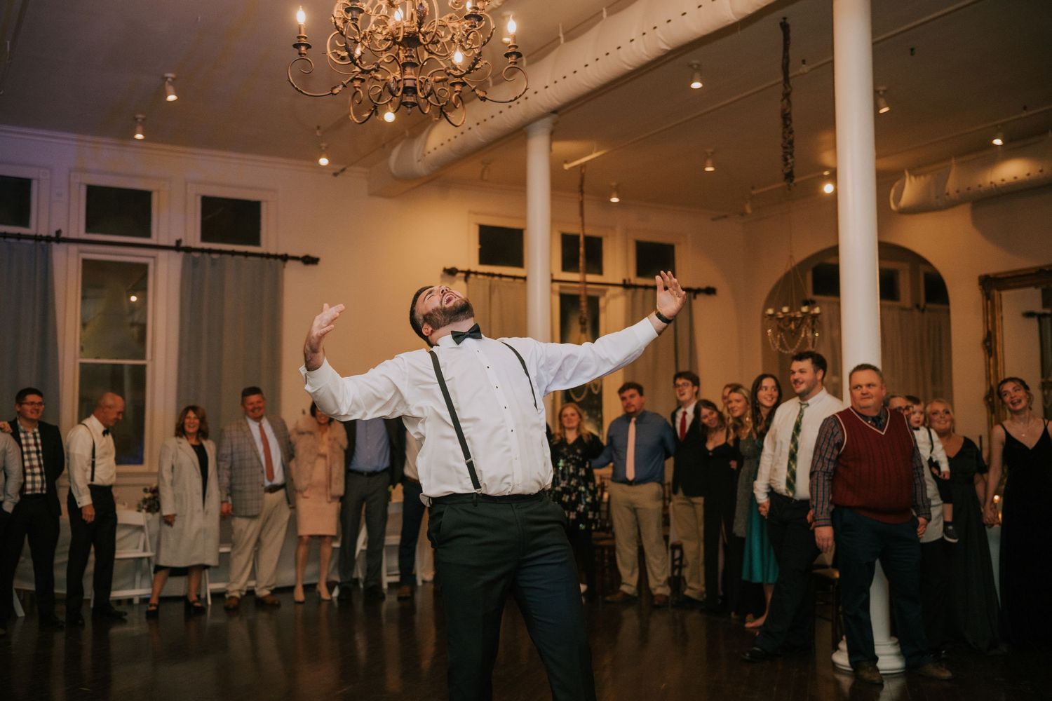 Wedding guest in suspenders dancing enthusiastically in an elegant ballroom with chandelier under guests' watchful eyes.