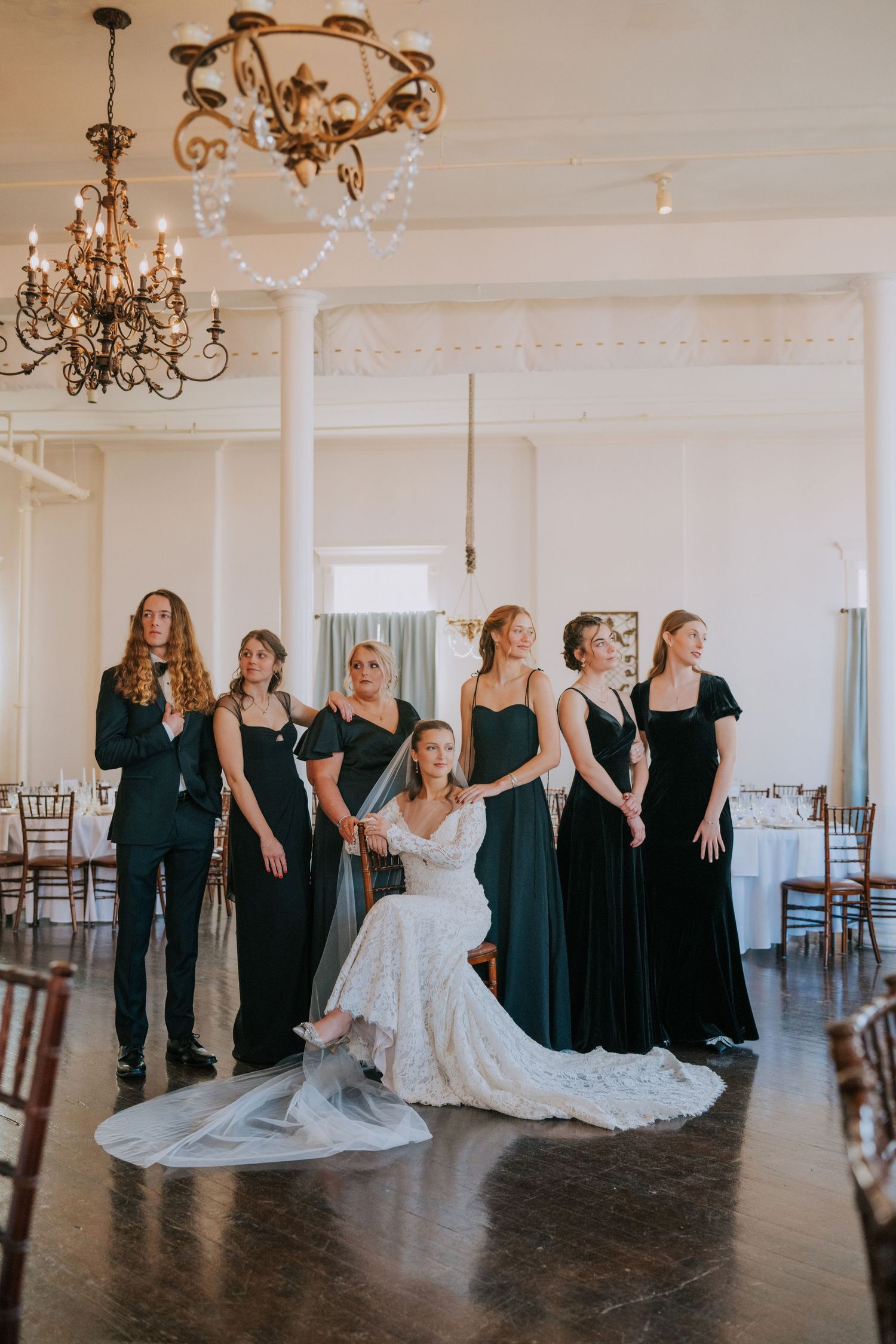 Wedding party poses in elegant reception venue with crystal chandeliers as bridesmaids wear dark gowns with white decor.