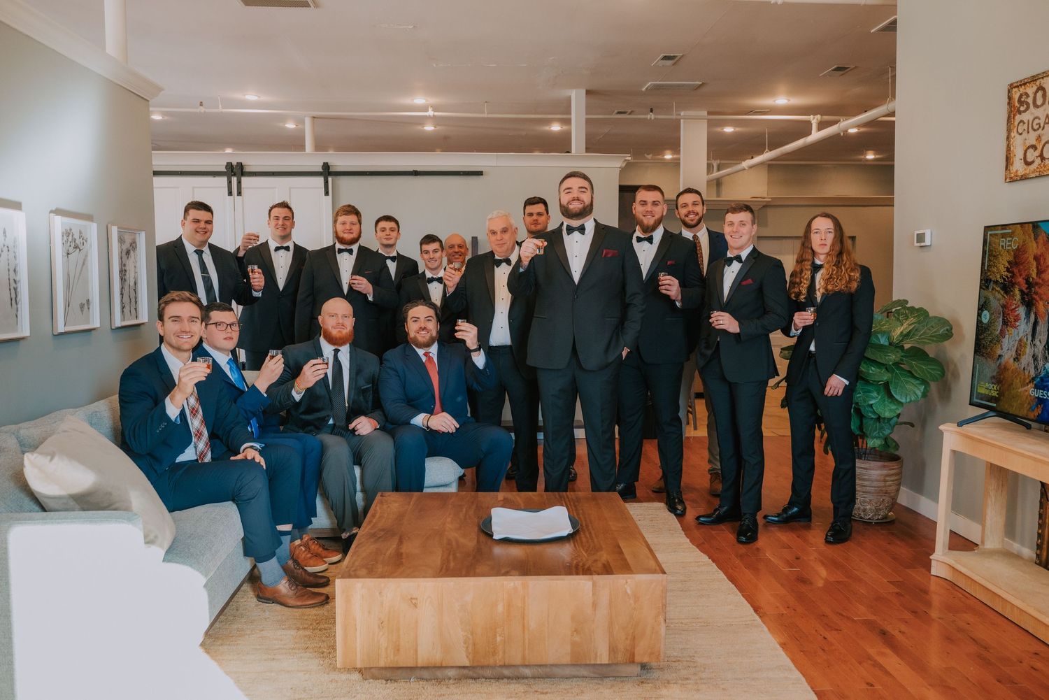 Wedding party group photo of groomsmen in formal suits gathered around a coffee table in an elegantly decorated room.