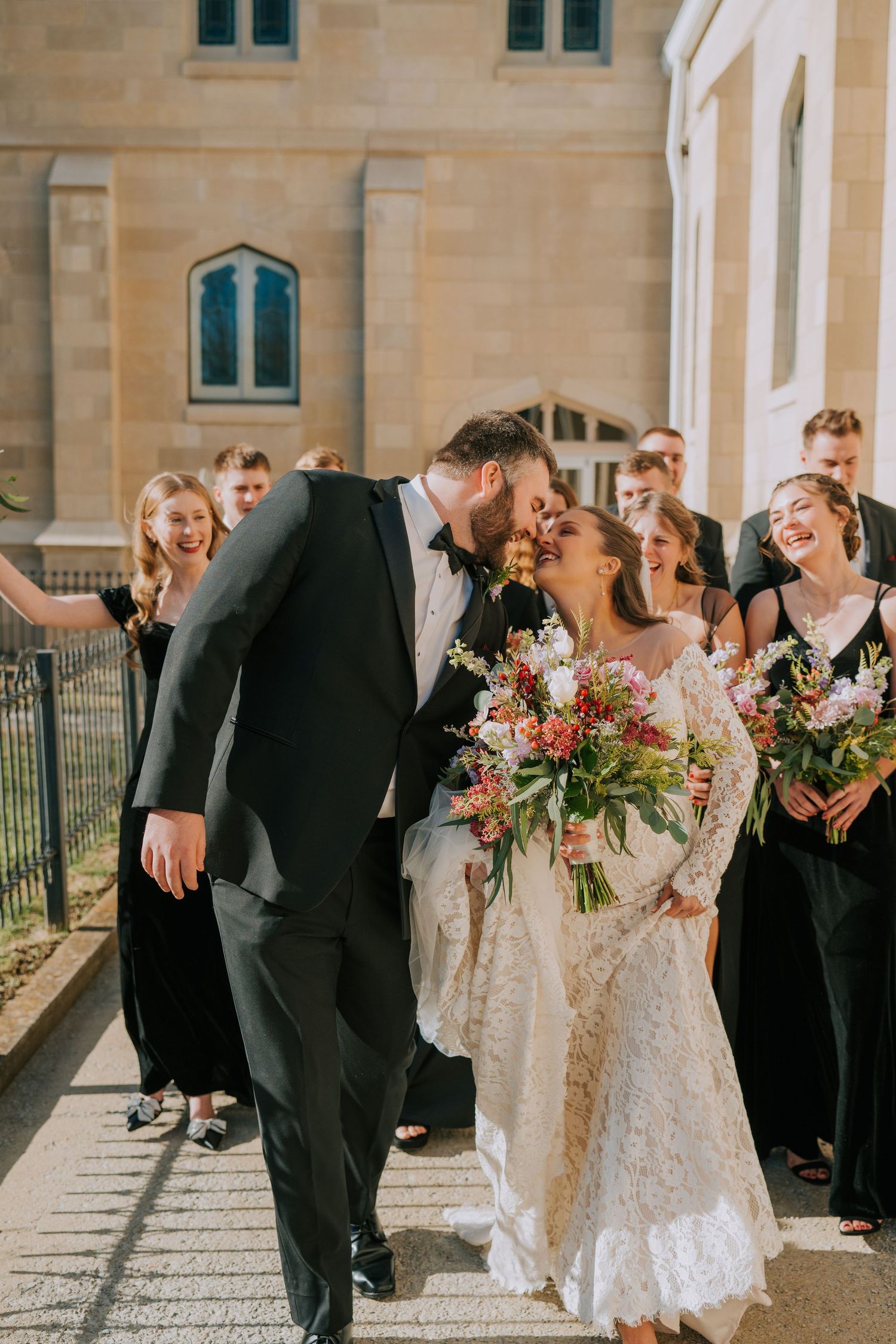 Wedding party laughing together outside church holding colorful floral bouquets.