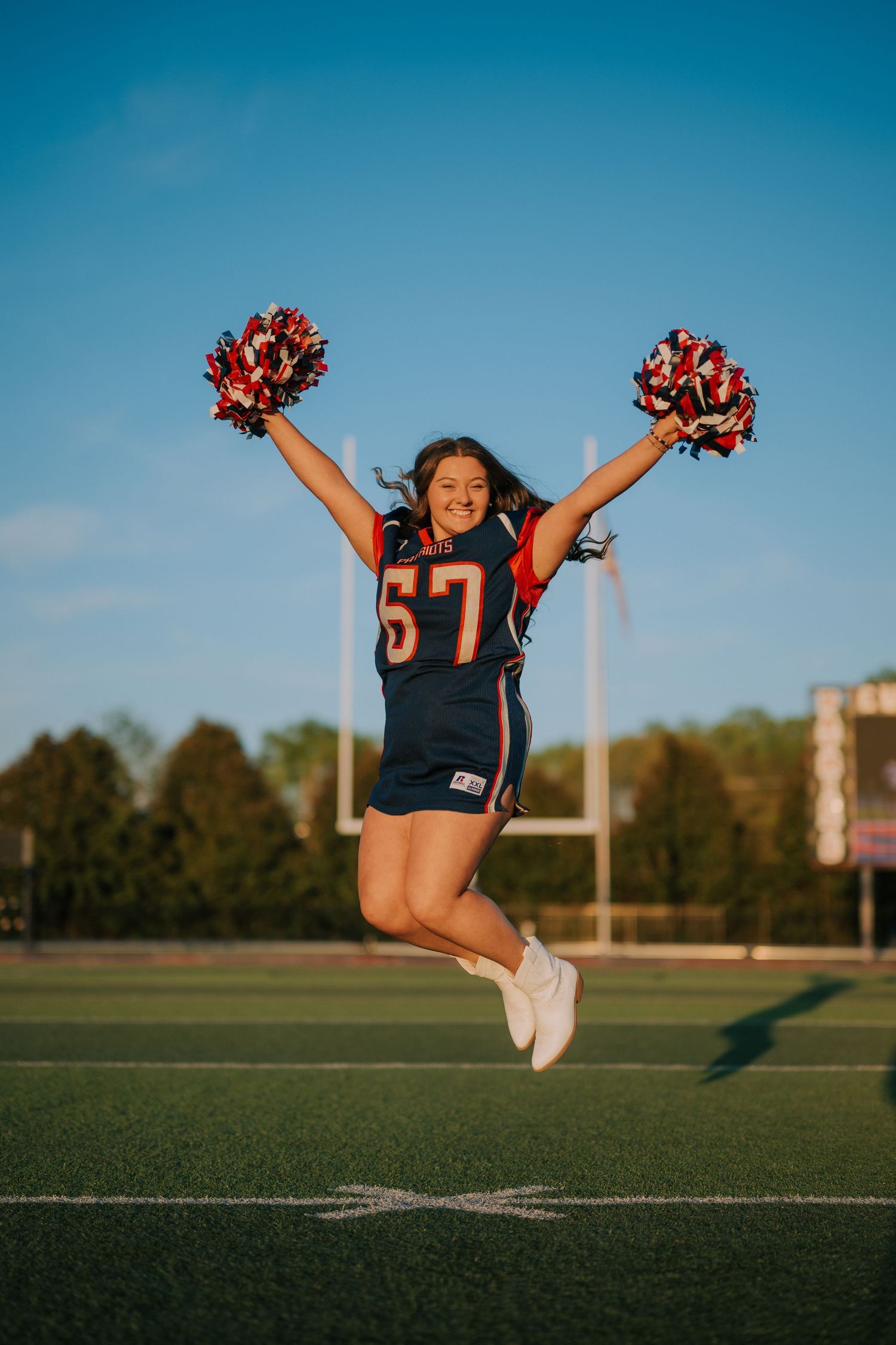 Cheerleader in sports jersey performs energetic jump with pom poms on football field at sunset.