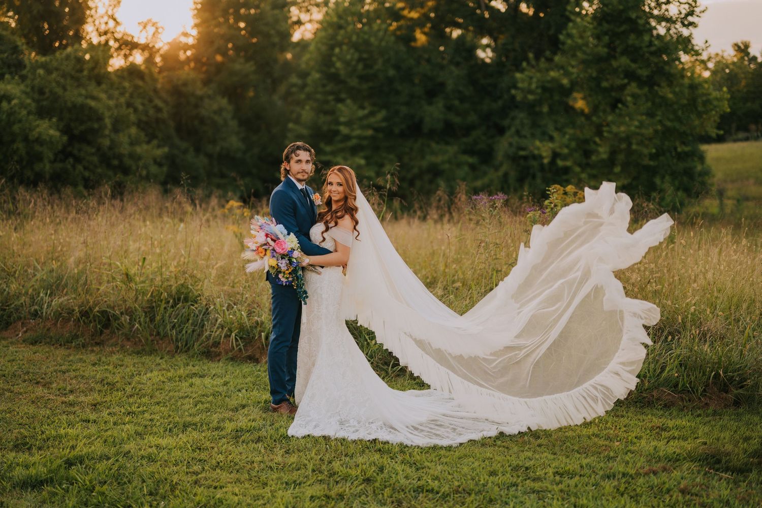 White wedding dress train flows dramatically across green grass at sunset.