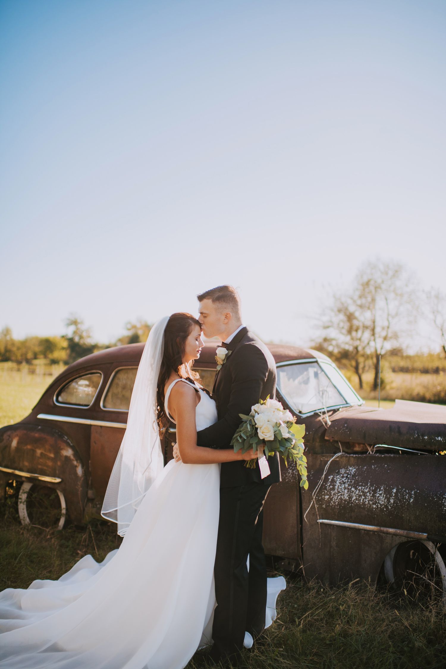 Newlyweds share an intimate moment near a rustic vintage car in a field.