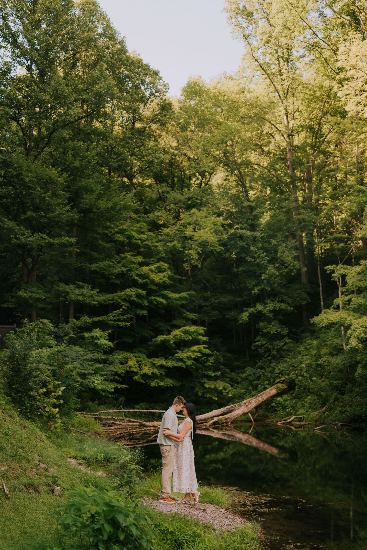 A couple stands together on a wooden bridge over a creek in a serene wooded setting.