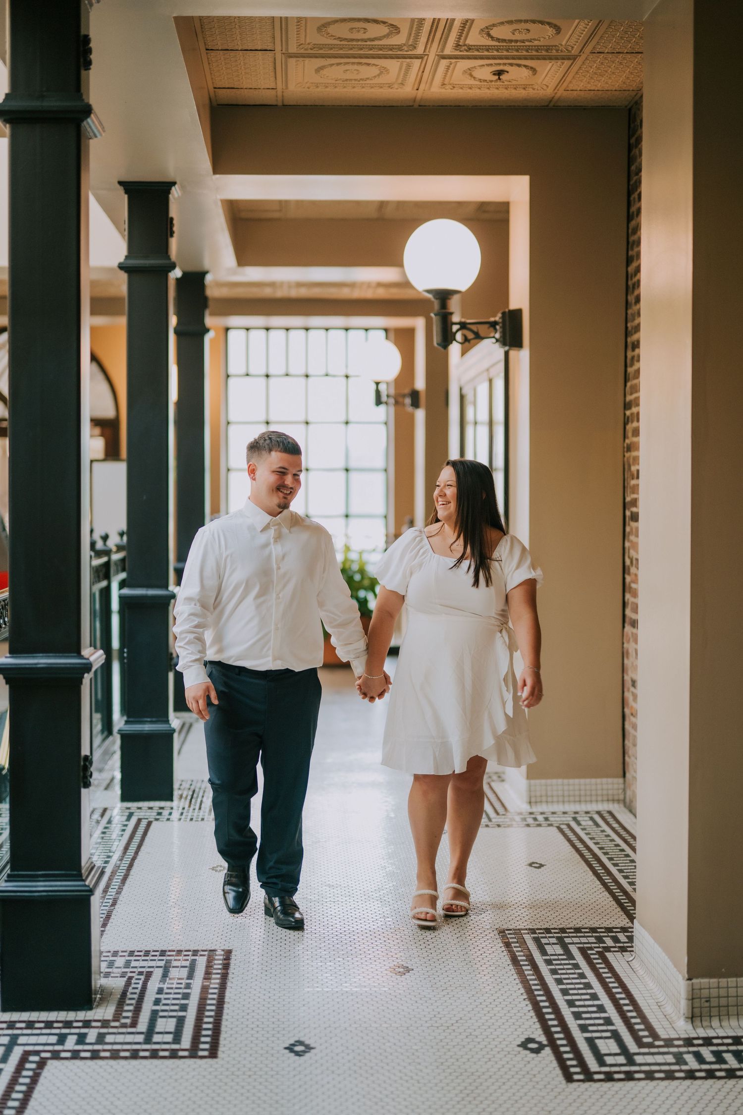 A couple walking through an elegant hallway with ornate tiled floors and decorative columns.