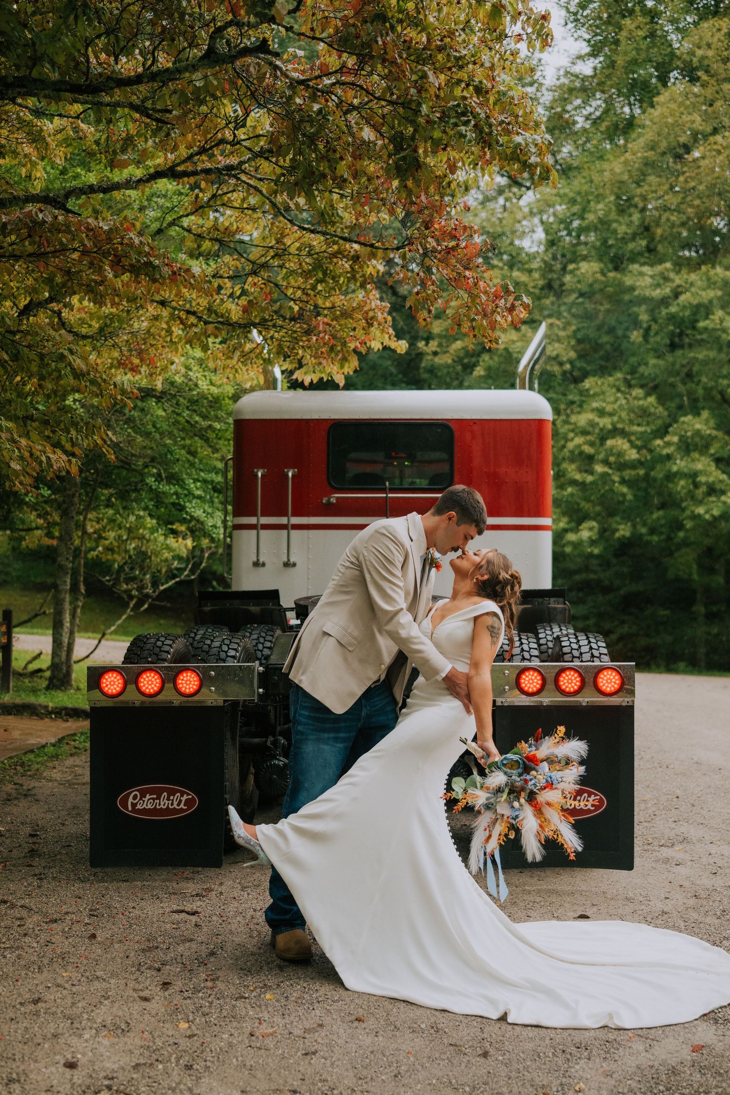 A romantic wedding couple shares a dip kiss in front of a vintage red truck surrounded by autumn trees.