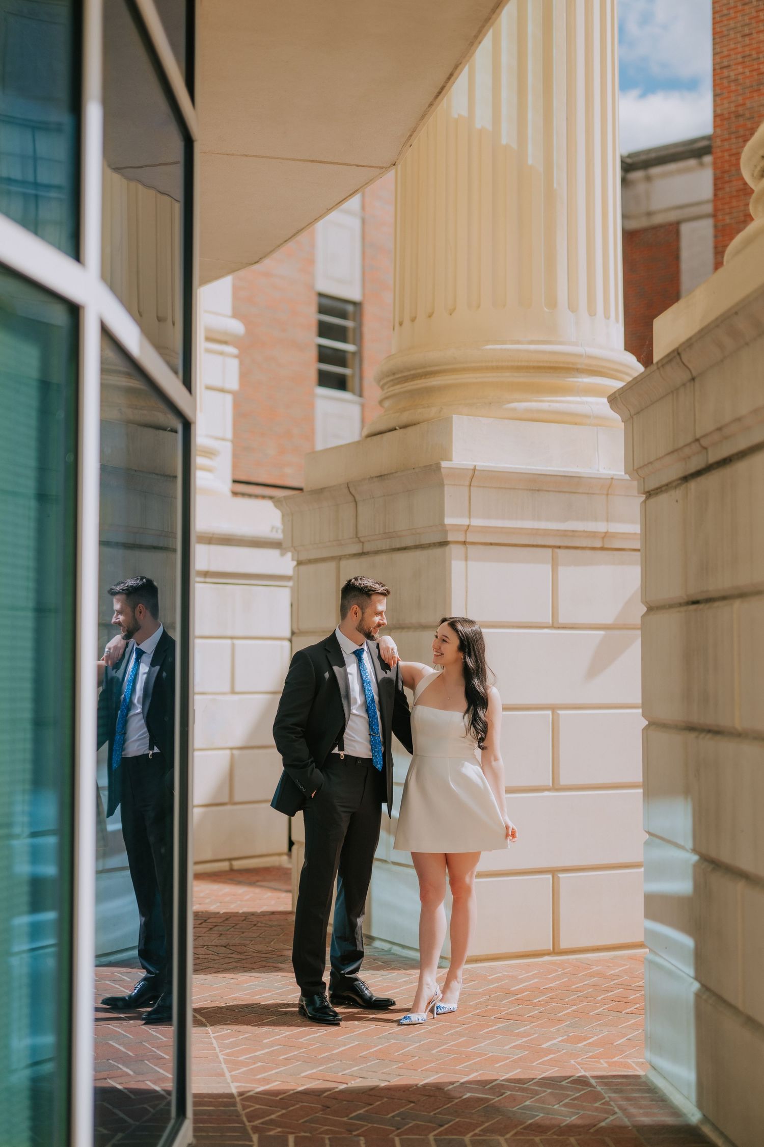 Elegant couple posing between white stone columns in formal attire.