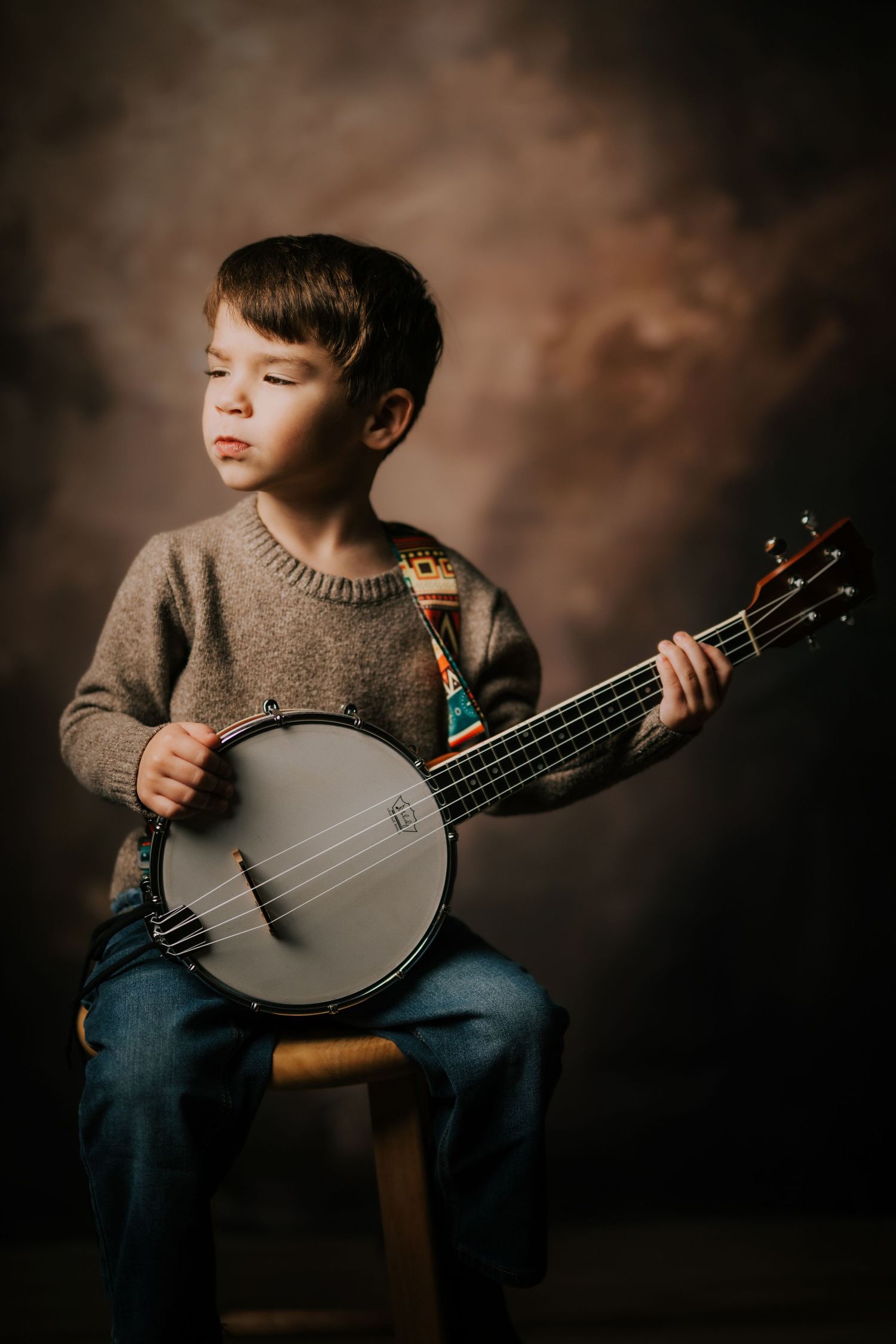 A child in a brown sweater and jeans plays a banjo in a moody studio setting with dramatic lighting.