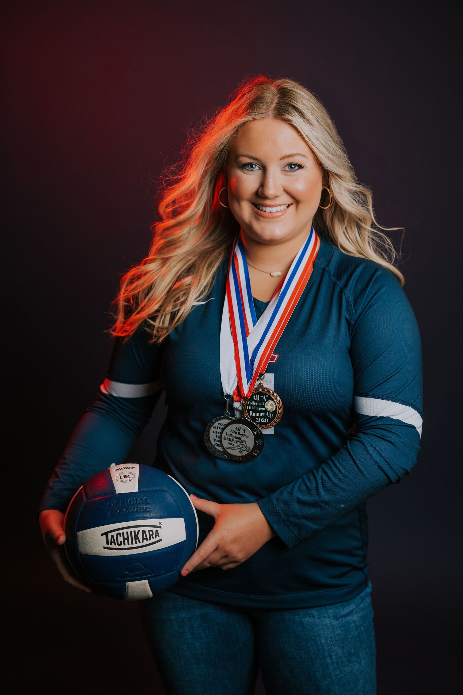 Athletic figure in blue top holds volleyball and wears medals against dark background with dramatic red lighting.