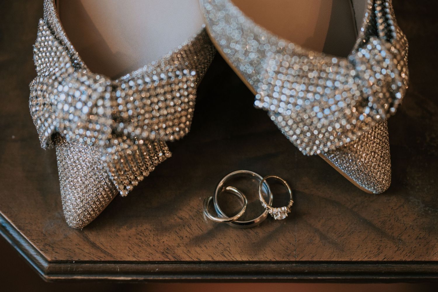 Sparkly crystal-embellished bridal shoes with bows displayed next to wedding rings on dark wooden surface.
