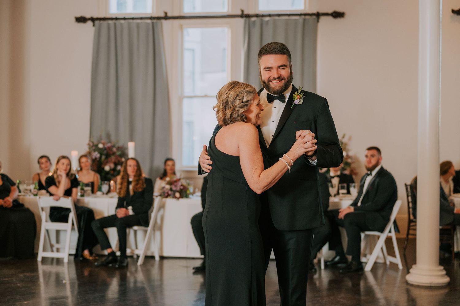 A couple shares an intimate dance at a wedding reception while guests watch from tables in an elegant white venue.