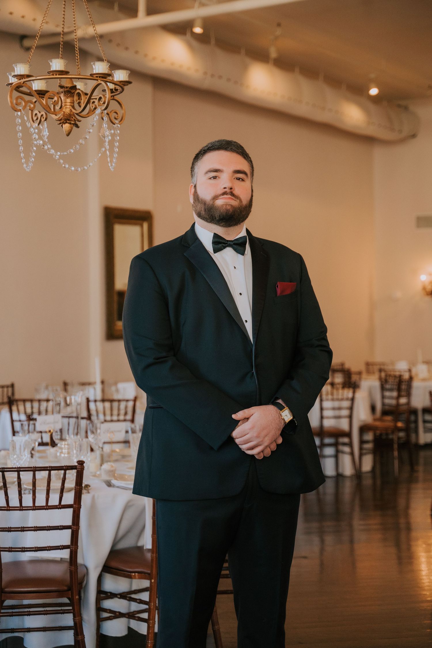 A groom in a classic black tuxedo stands in an elegant wedding venue with chandelier lighting and Chiavari chairs.