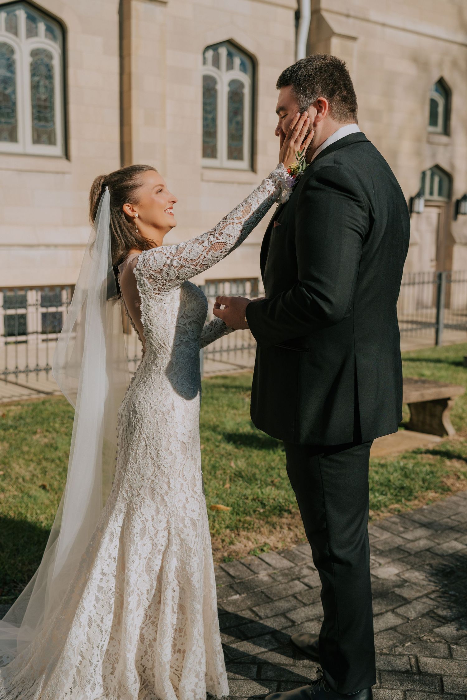 A bride in a long-sleeved lace wedding dress touches her groom's face tenderly outside a church building.