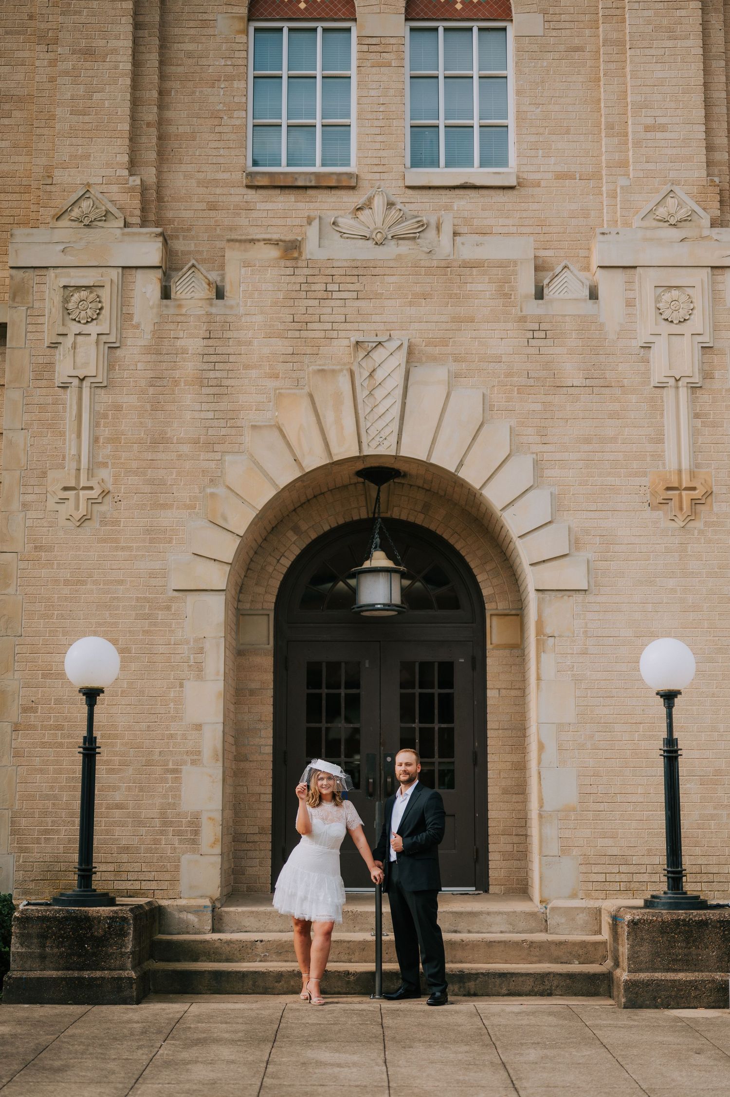 A couple poses outside a grand historic brick building with arched doorways and decorative architecture.