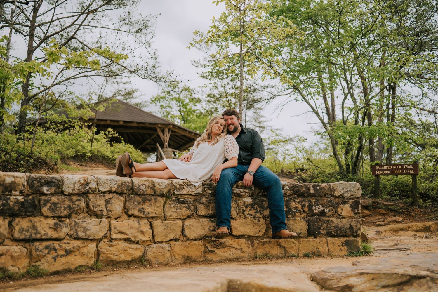 Couple sitting on rustic stone wall surrounded by trees during outdoor photoshoot.