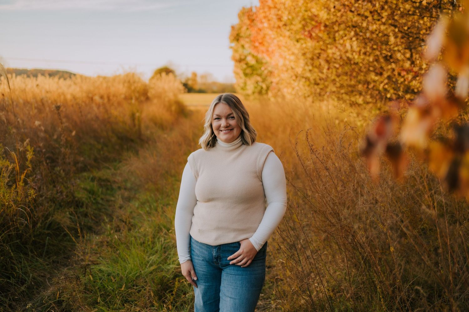 Woman in cream sweater smiling while standing on nature path with autumn trees.