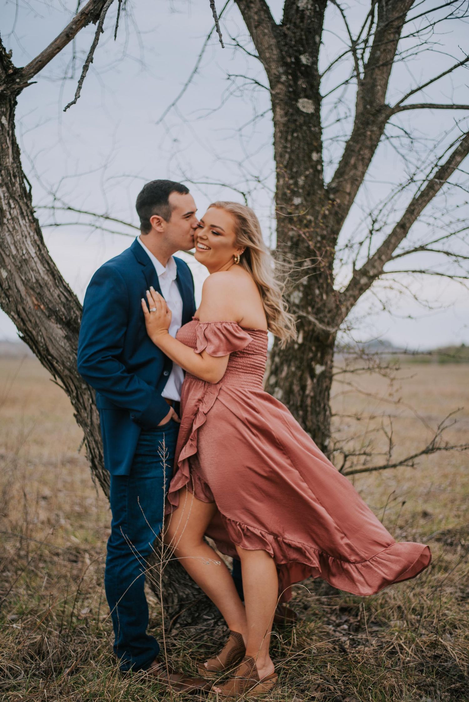 A romantic couple embraces under bare trees in a rustic outdoor setting.
