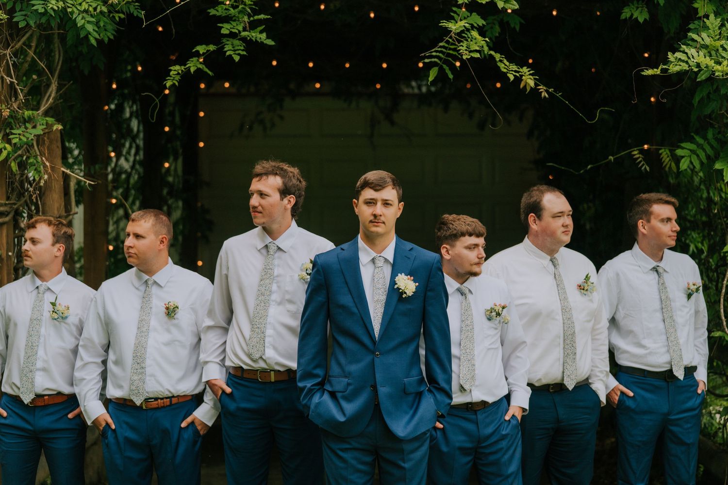 Wedding party in navy suits and white shirts lined up outdoors under string lights.