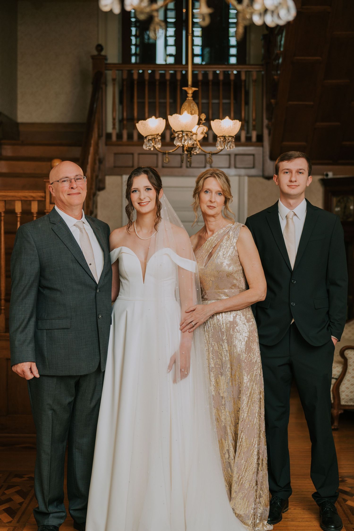 Formal wedding party portrait with family members in elegant attire standing beneath a chandelier in a classic venue.
