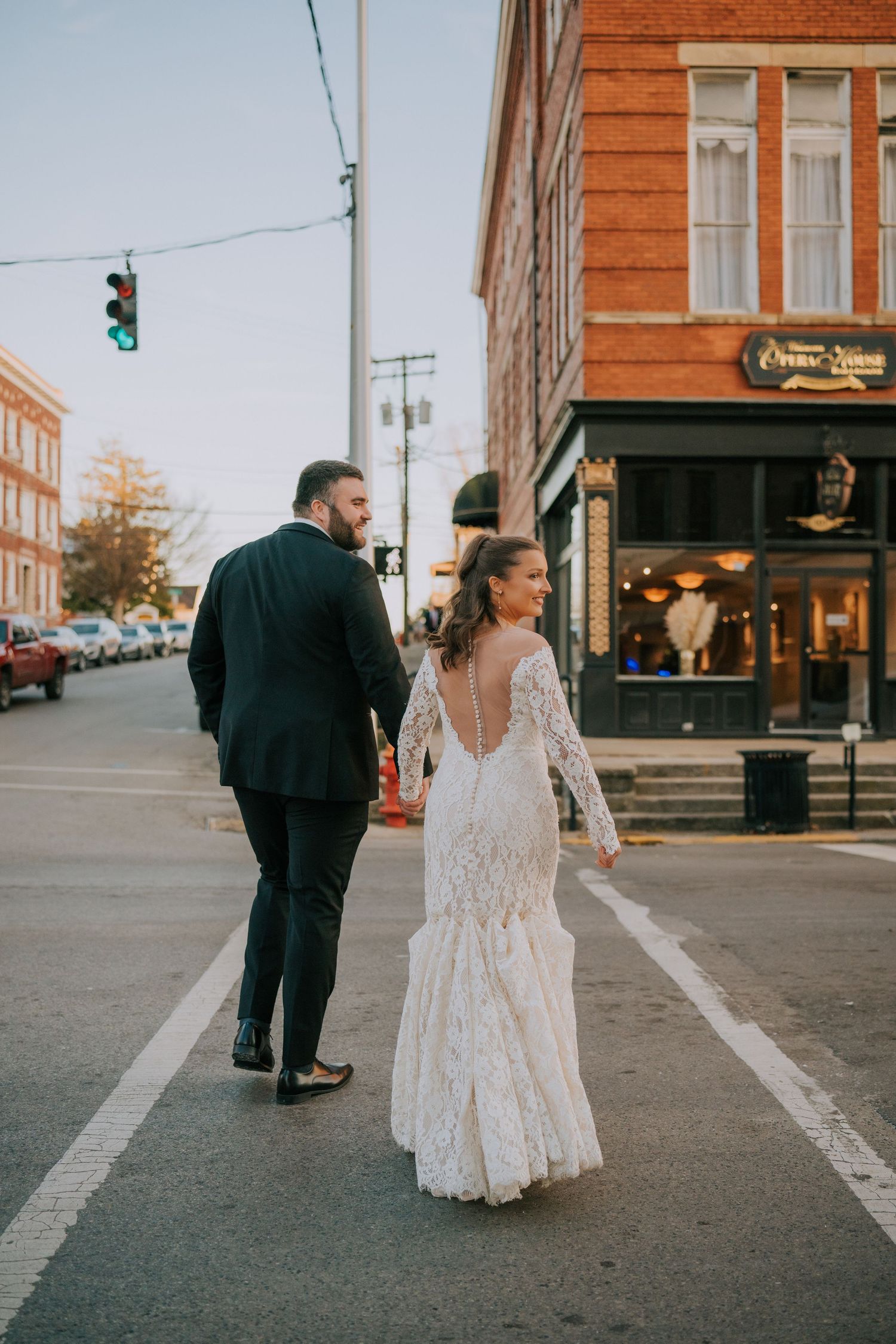 A couple in wedding attire walks along a city street with brick buildings in the background.