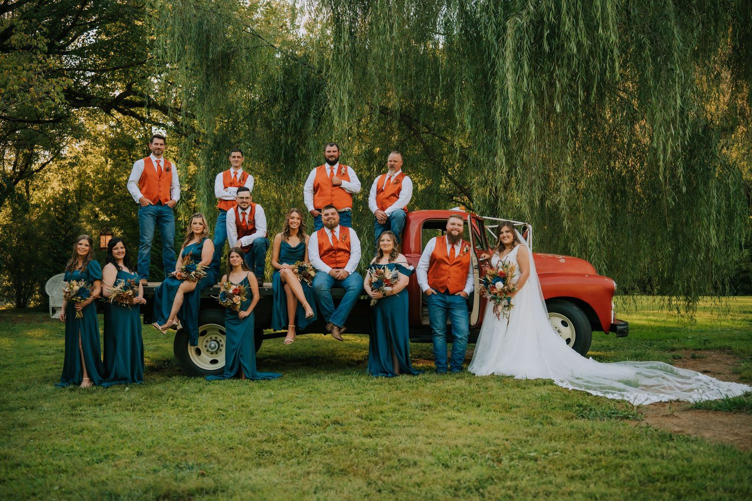 A wedding party poses with a vintage red truck under willow trees.