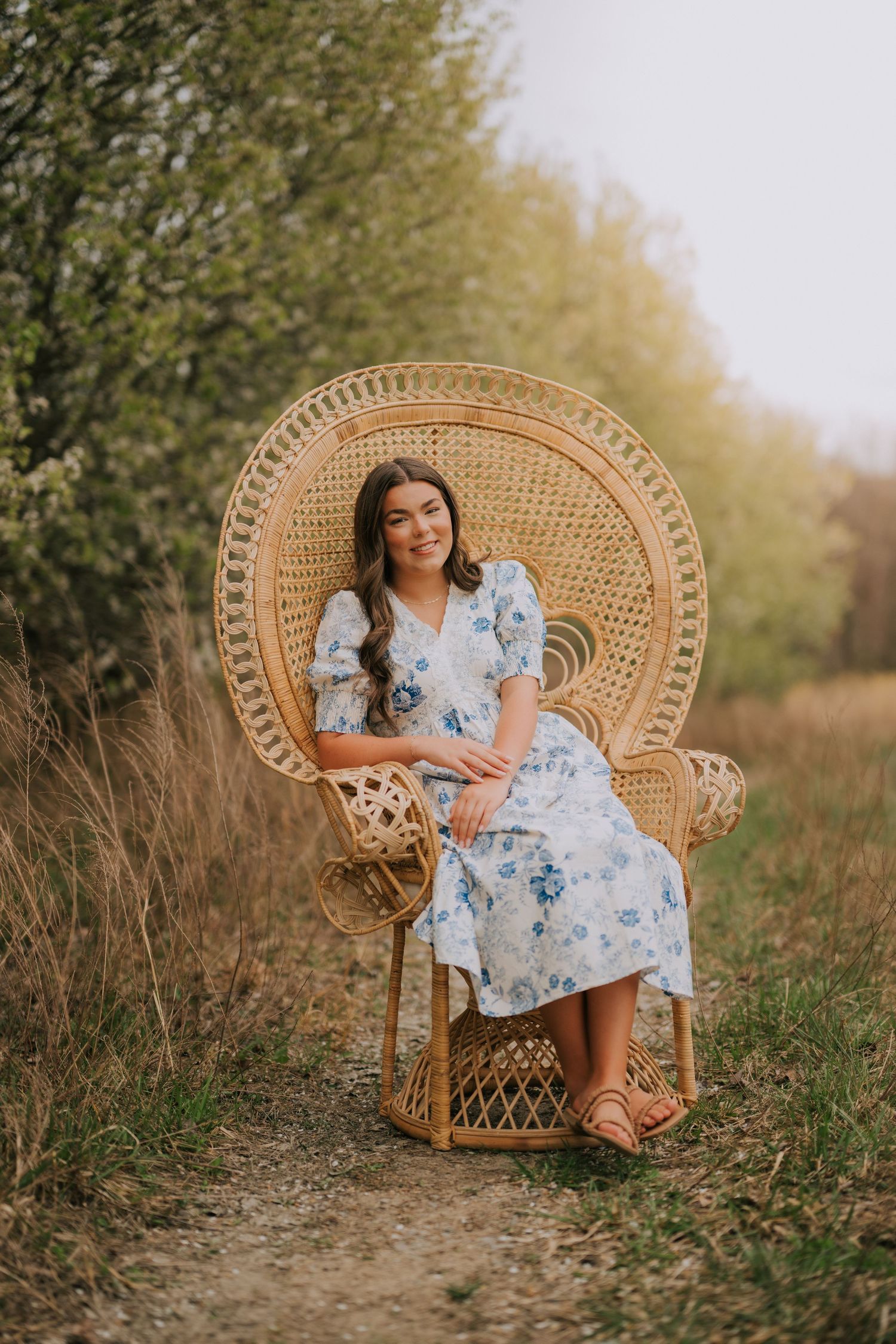 Natural portrait session featuring wicker peacock chair in golden field setting.
