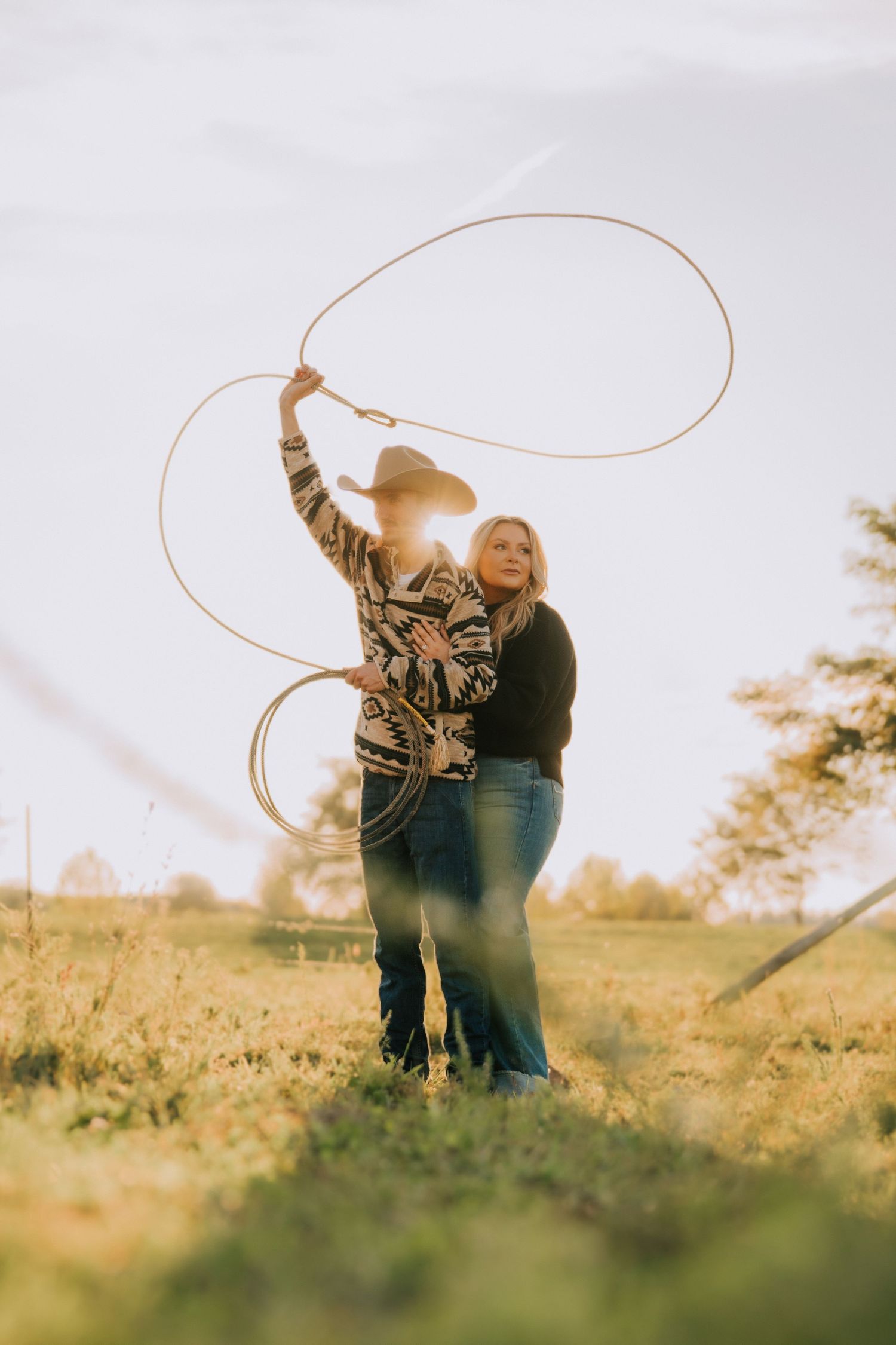 Couple practices rope tricks together in sunlit field wearing western clothing.