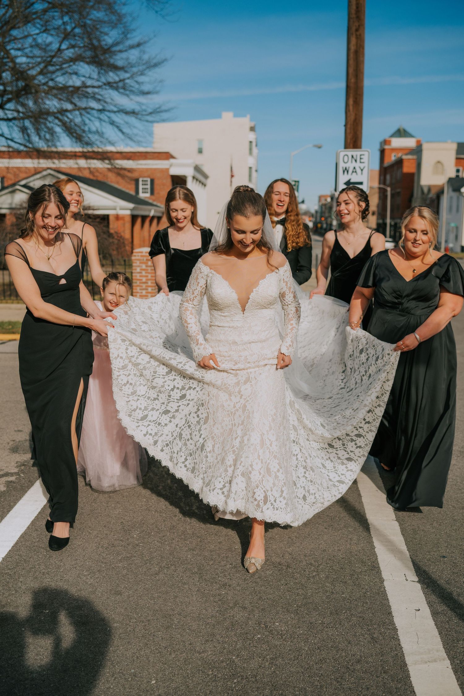 A bride in a white lace wedding dress poses with bridesmaids in black dresses on a sunny day outdoors.