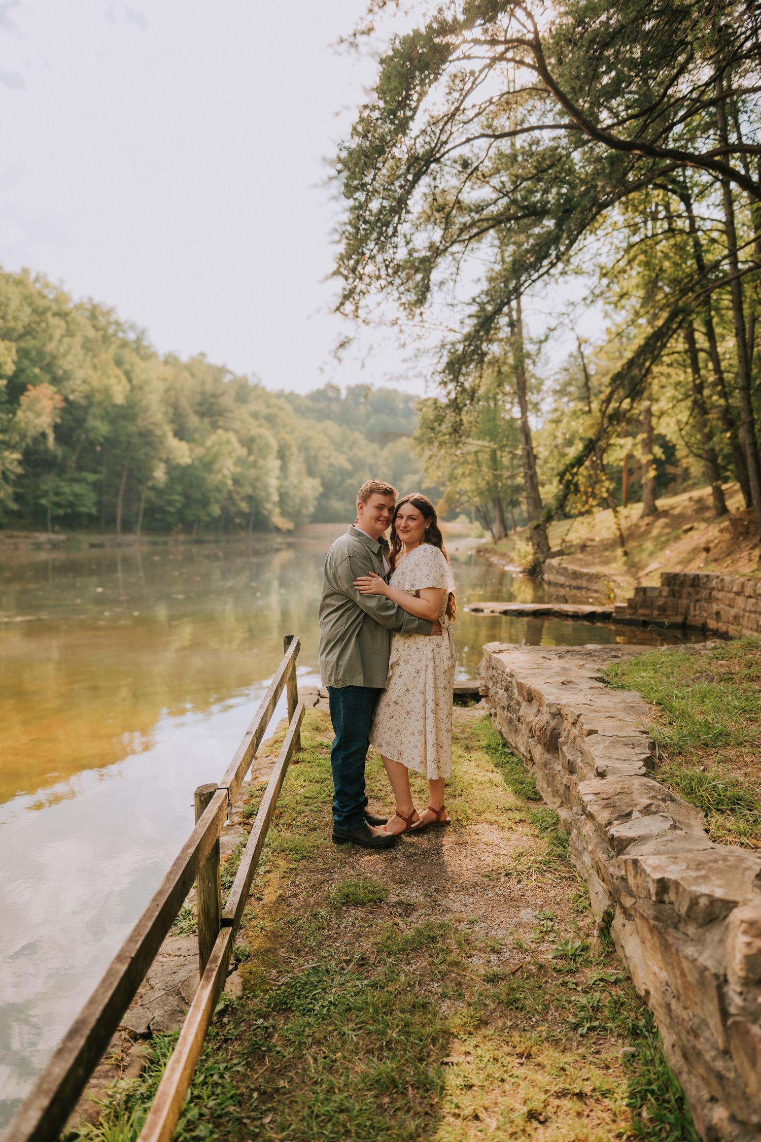 A couple enjoys a romantic moment along a riverside path with stone walls and autumn trees.