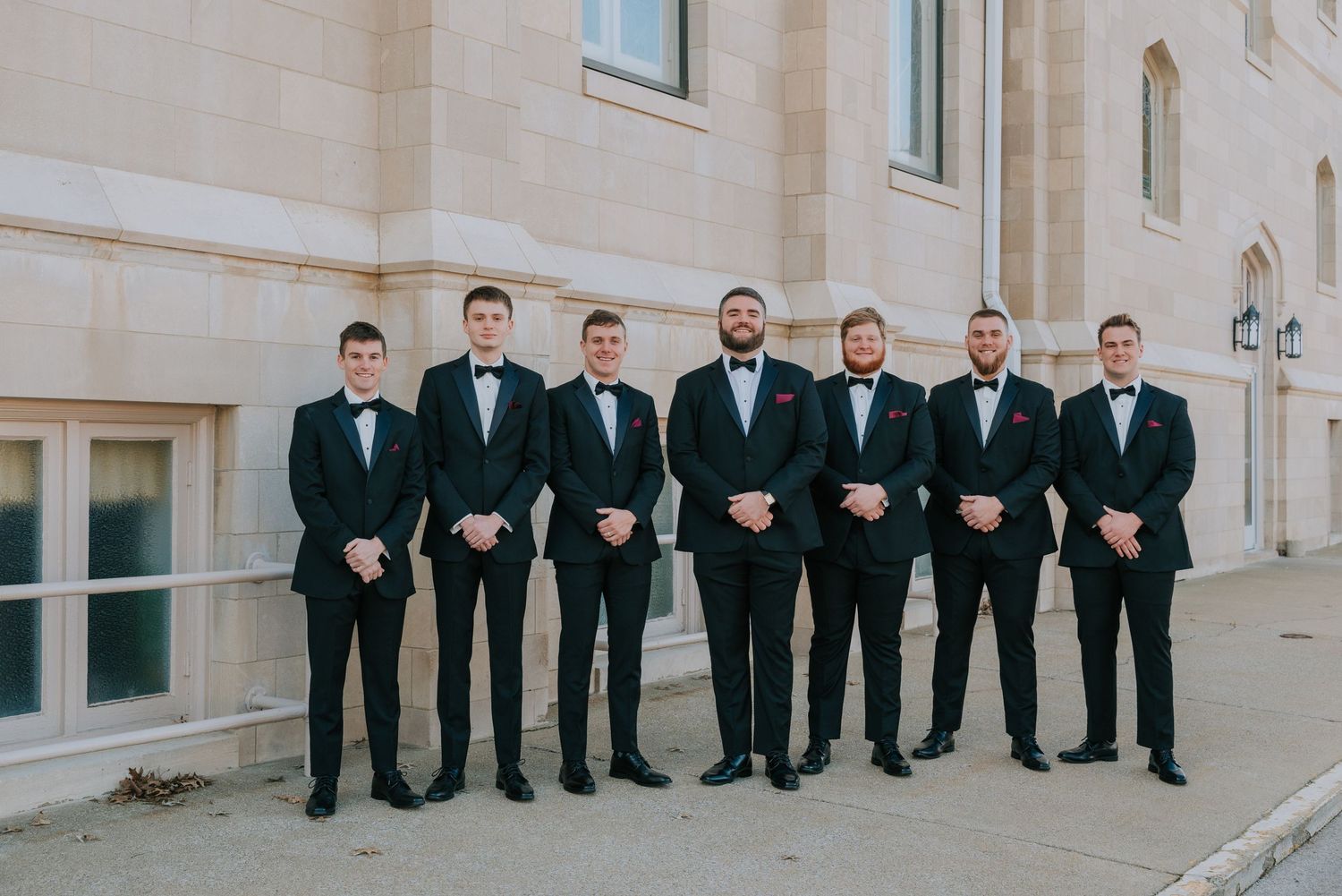 A group of groomsmen in matching black tuxedos and bow ties stand in a line against a light stone building exterior.