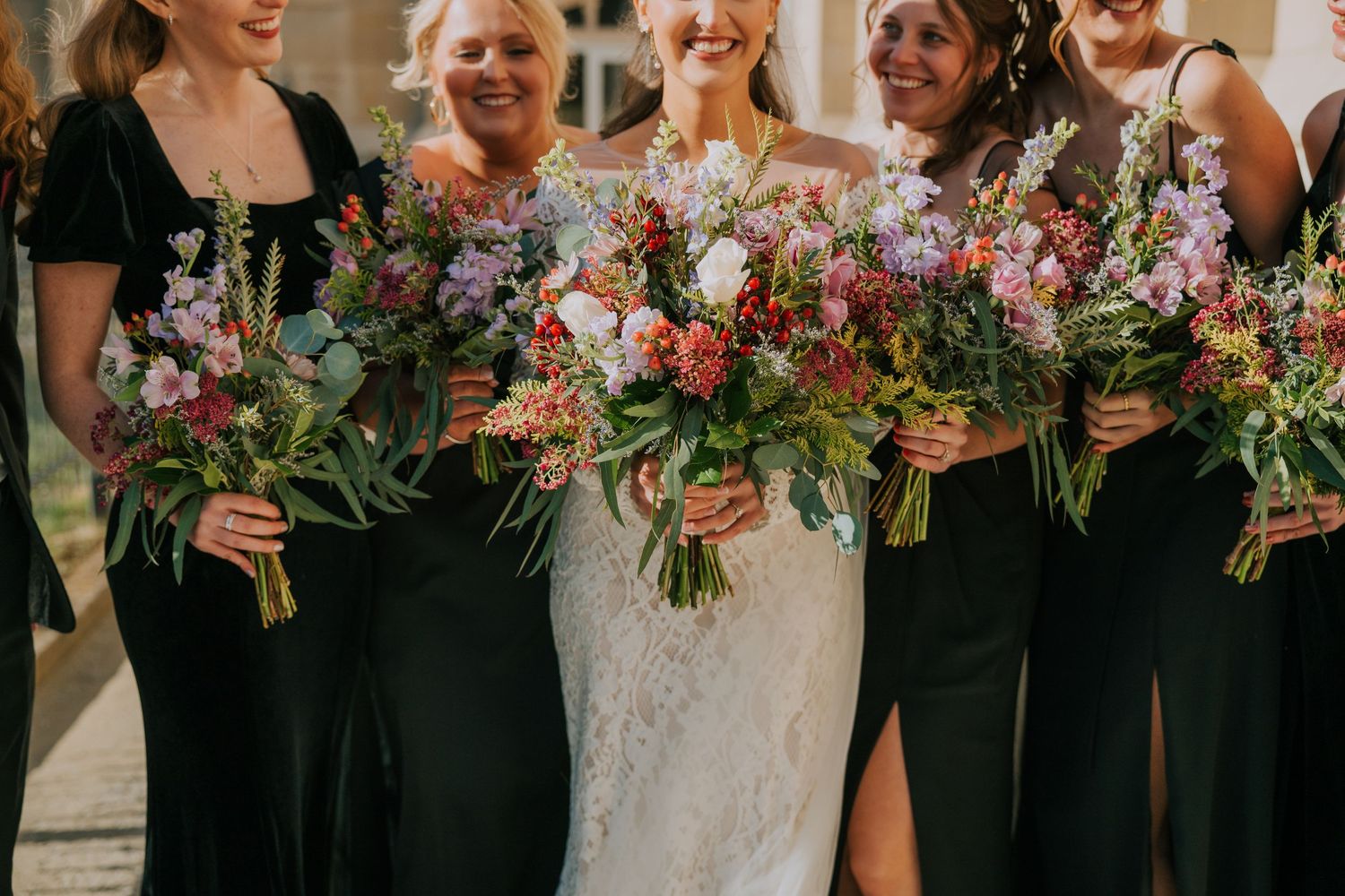 A bridal party wearing black dresses and carrying vibrant wildflower bouquets stands together at a wedding celebration.