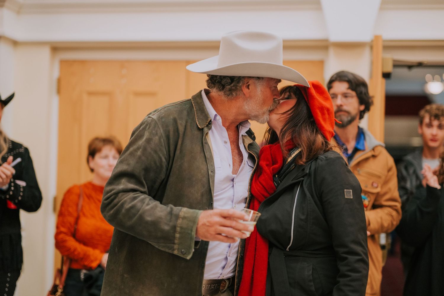 A couple shares a tender moment at what appears to be an outdoor gathering with people watching in the background.