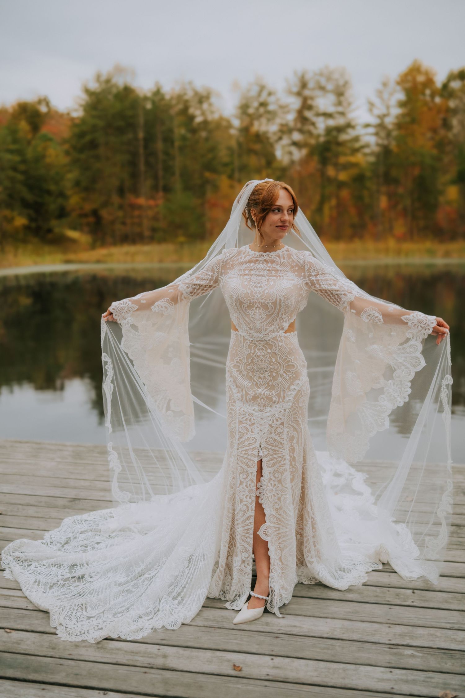 Bride in lace gown displays flowing train on wooden dock by autumn lake.