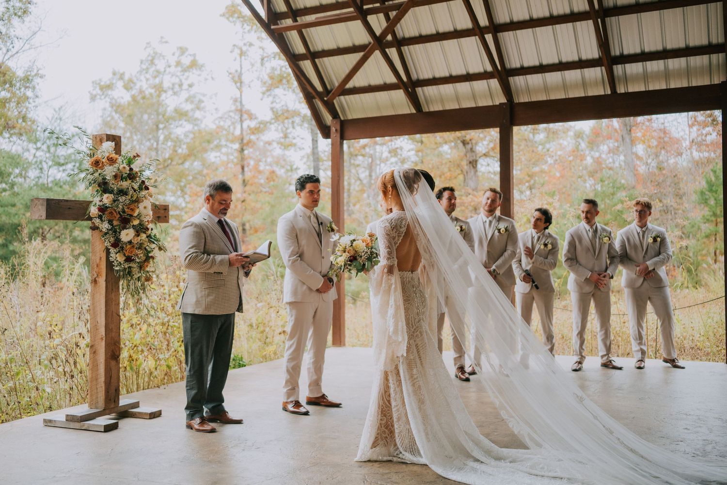 A wedding ceremony takes place in a rustic outdoor venue with autumn trees visible in the background.