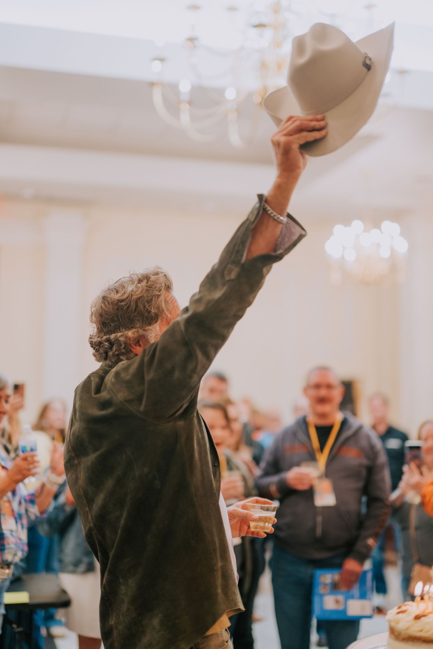 A person raises a cowboy hat in celebration while others cheer in the background at an indoor event.