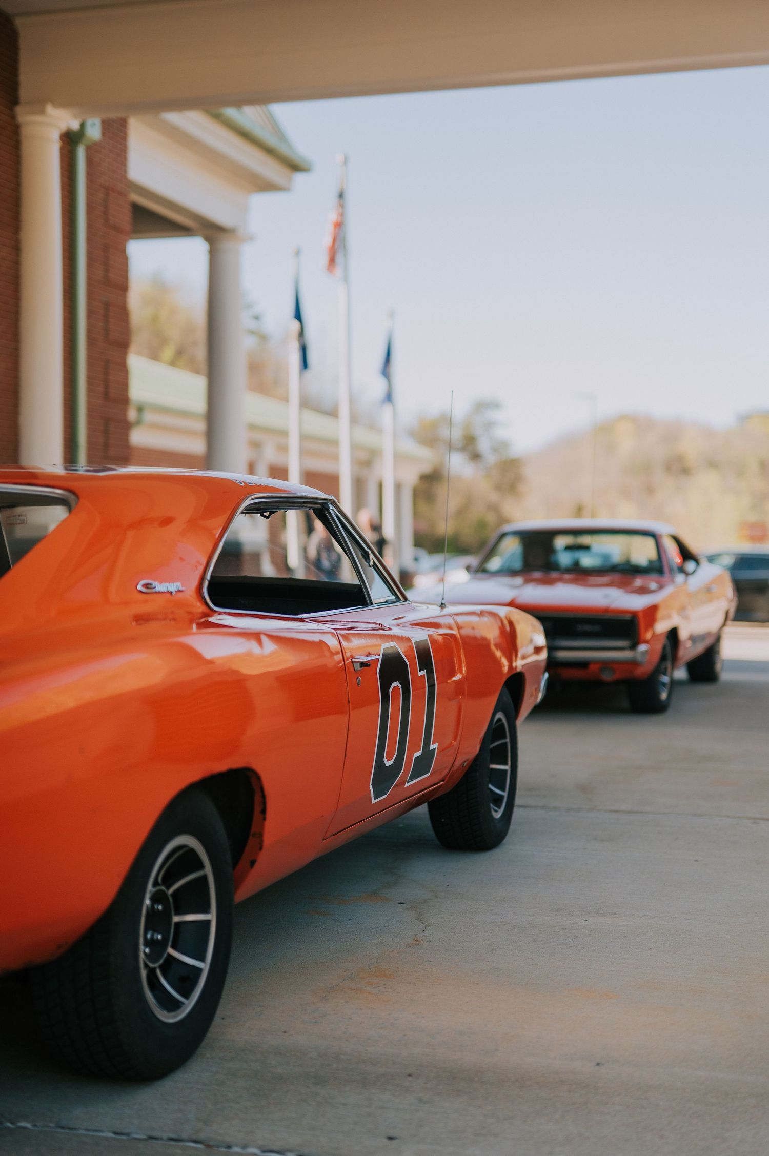 Two classic orange muscle cars, including the iconic General Lee Dodge Charger from Dukes of Hazzard, parked at a building entrance.