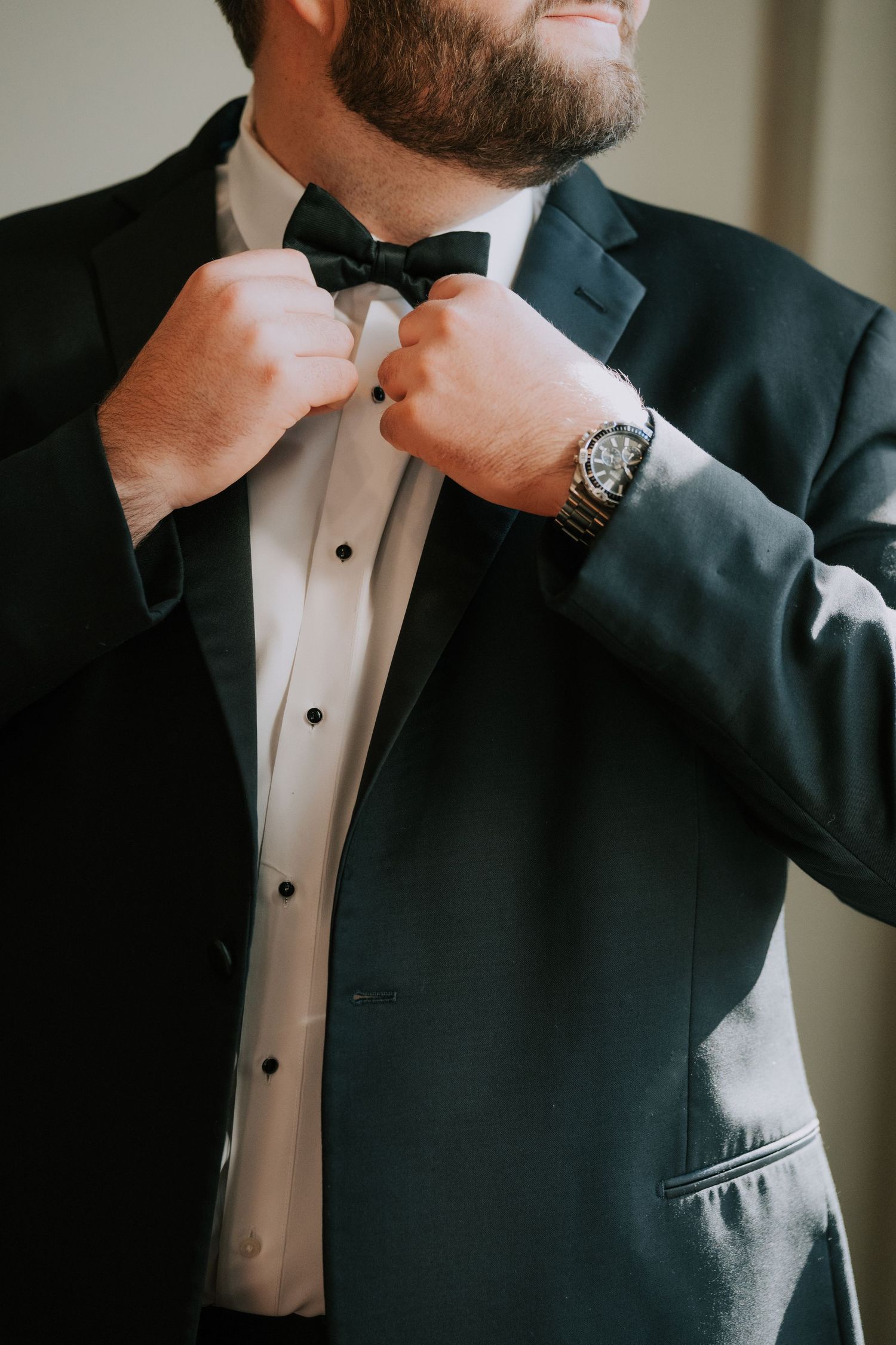Close up detail of hands adjusting a black bow tie on a formal tuxedo with dress shirt and watch visible.