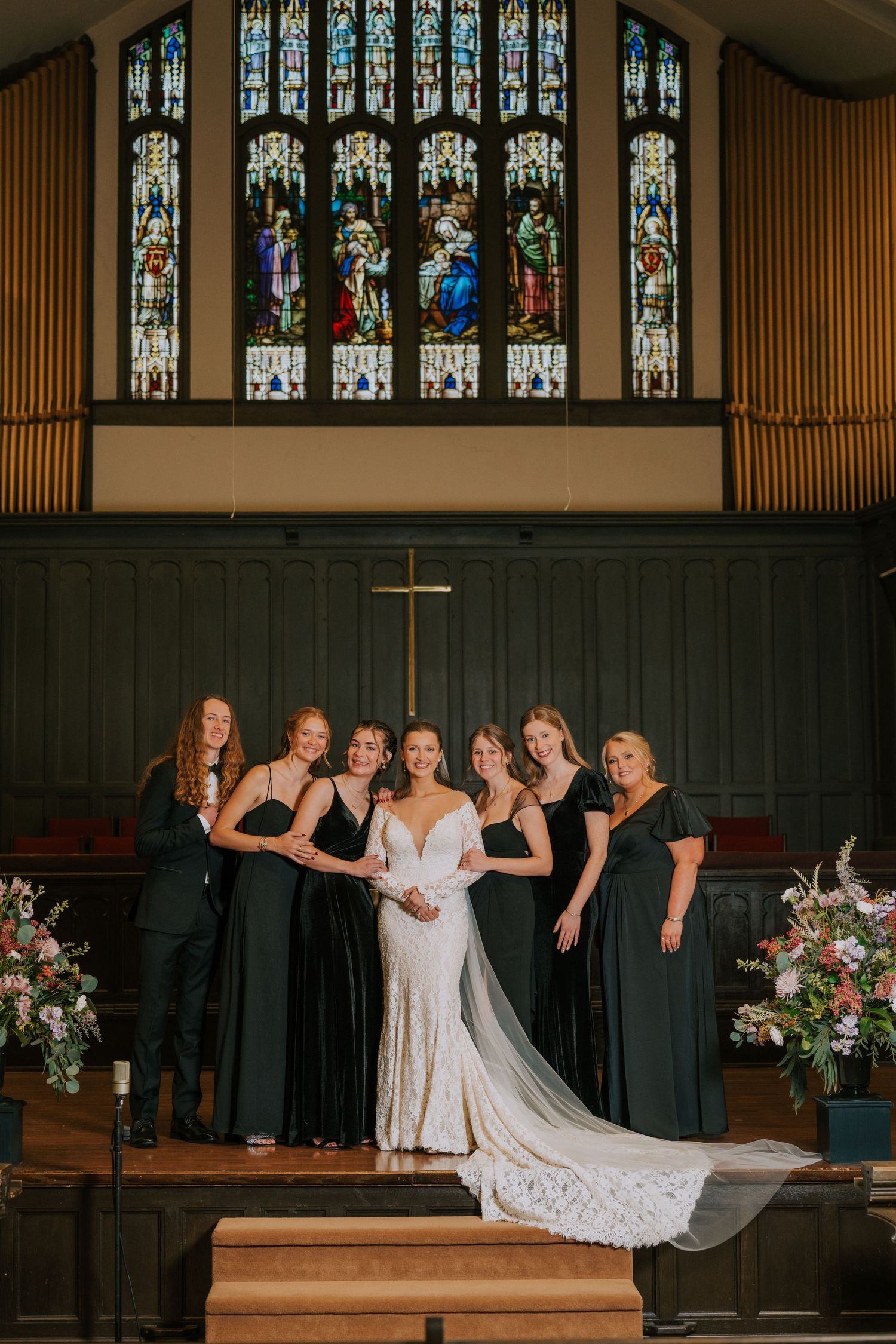 A bridal party poses in a church sanctuary with beautiful stained glass windows and a wooden cross behind them.